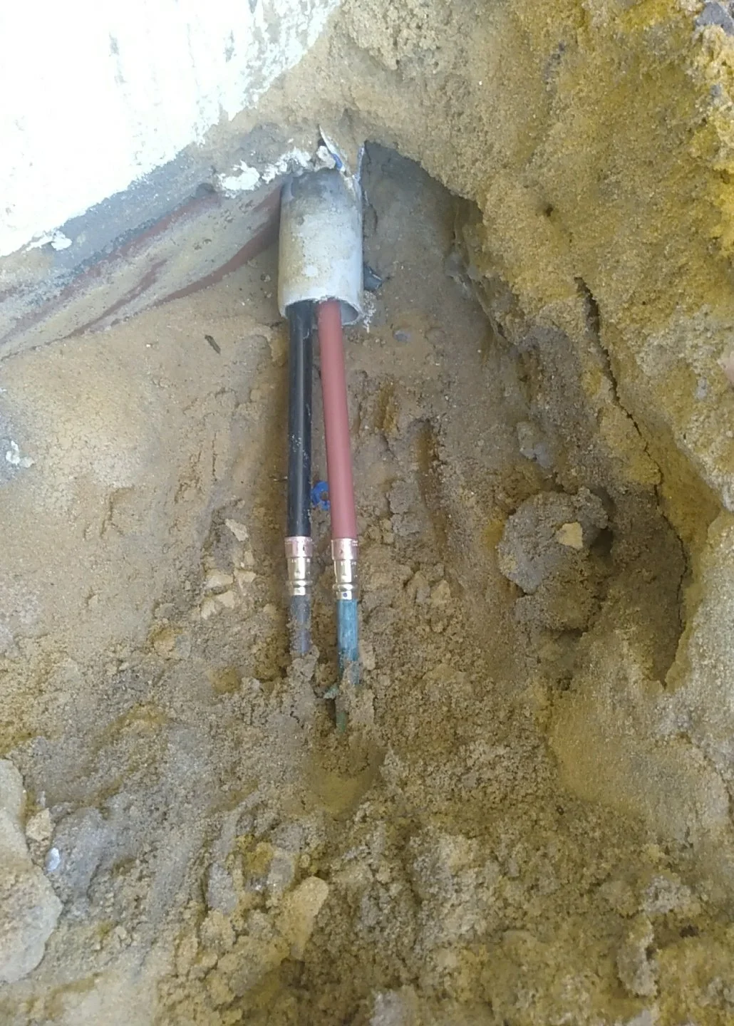 Installation of electrical or plumbing pipes emerging from a wall with exposed yellowish soil and drywall in the background.