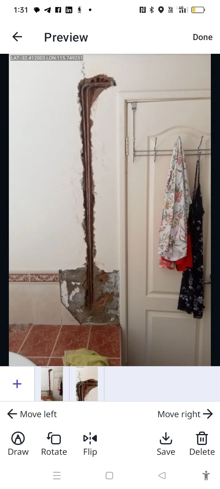 A bathroom wall with exposed pipes and damaged drywall near a door, with clothes hanging on hooks.