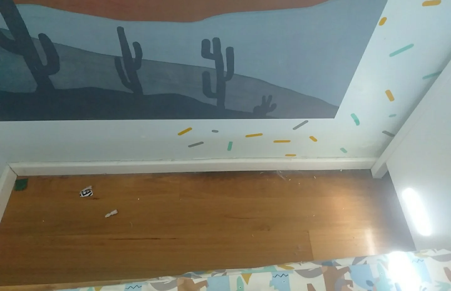 Corner of a room with a wooden floor, a colorful patterned curtain at the bottom, a white baseboard, and a painted wall with a desert scene featuring cacti and a hand, decorated with scattered multicolored sprinkles.