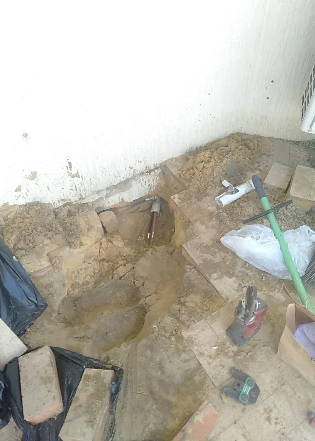 Underground plumbing installation site with exposed pipes, tools, and dirt near a white wall.
