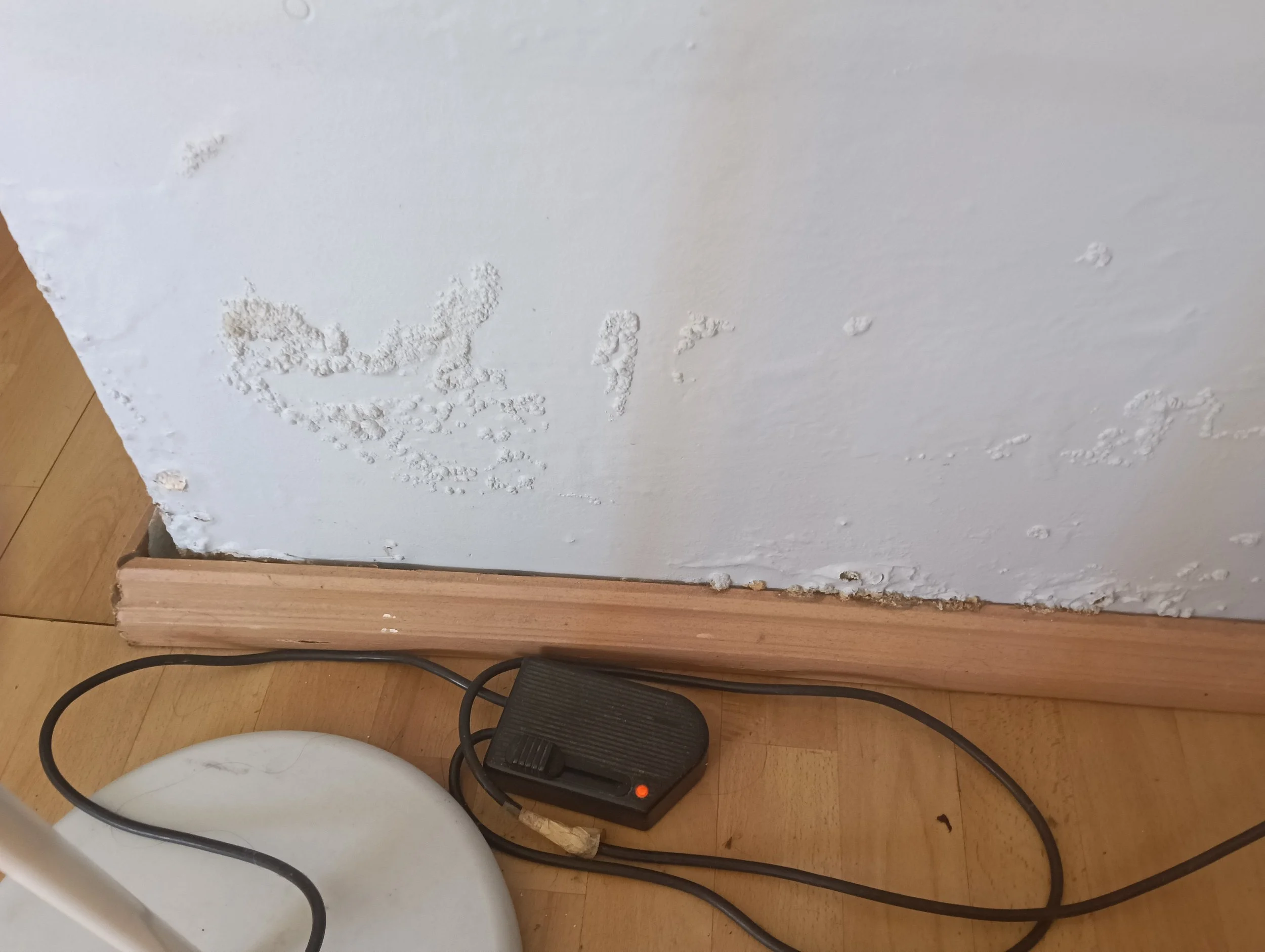 Interior wall with peeling paint and damaged drywall near the baseboard, with an electrical power strip on a wooden floor.