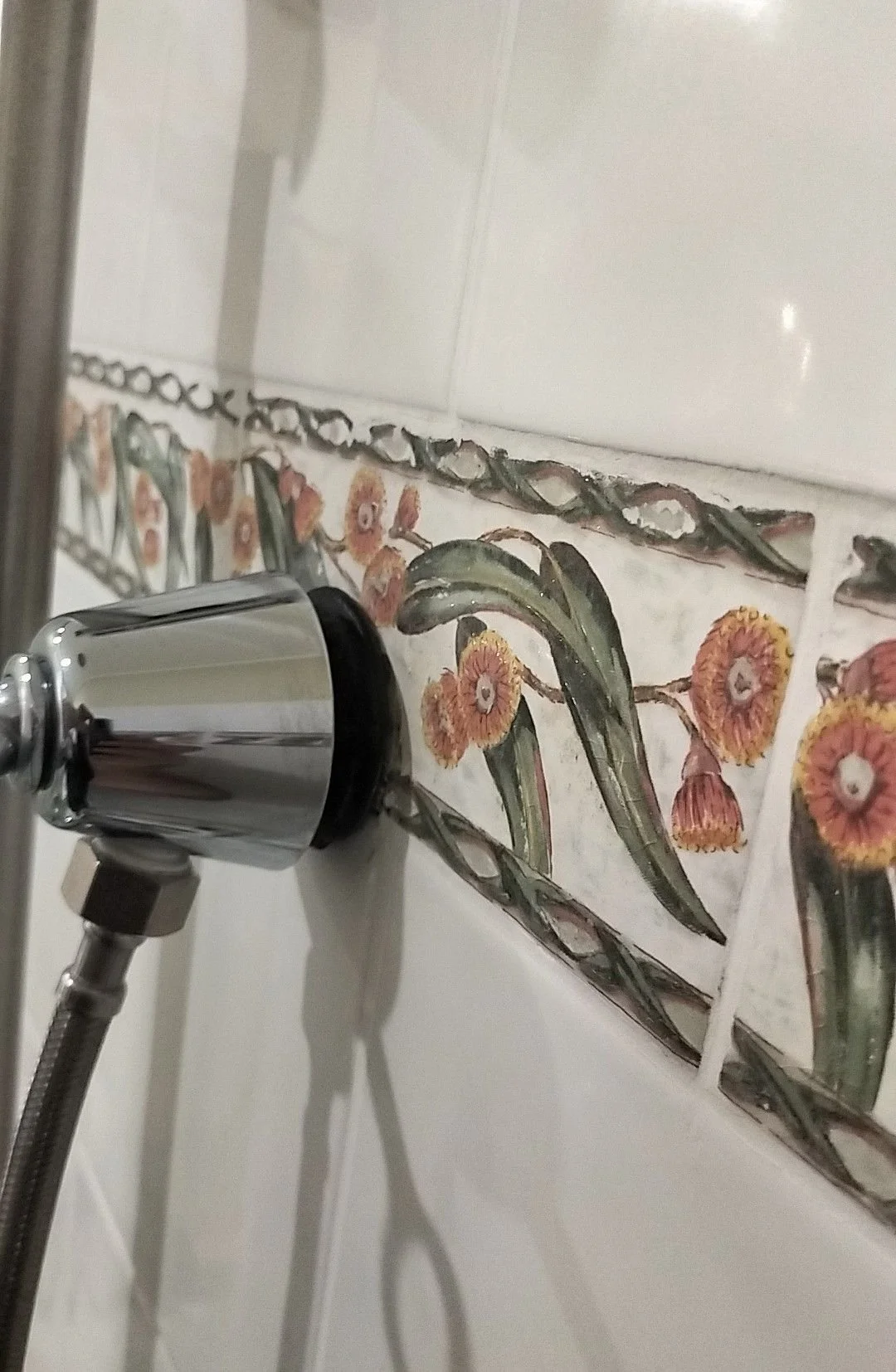 Close-up of a chrome shower handle mounted on a white tiled bathroom wall with decorative floral and leaf pattern tiles.