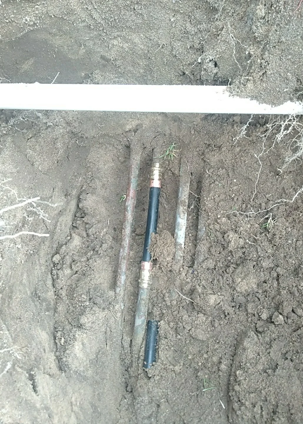 Underground plumbing pipes installed in a trench with soil surrounding them, and a white pipe placed on the ground above.