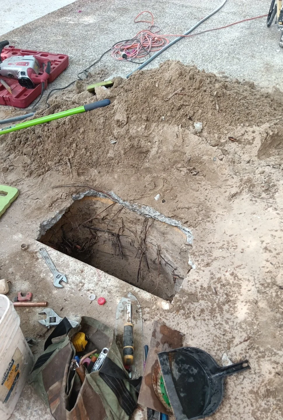 Open excavated hole in a concrete floor showing underground pipes and wires, surrounded by construction tools and equipment.