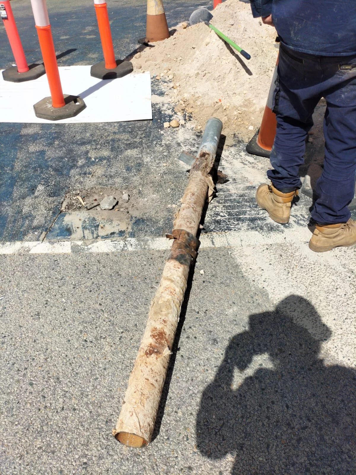 A construction worker standing next to a damaged utility pole on the ground at a construction site with safety cones and sand pile in the background.