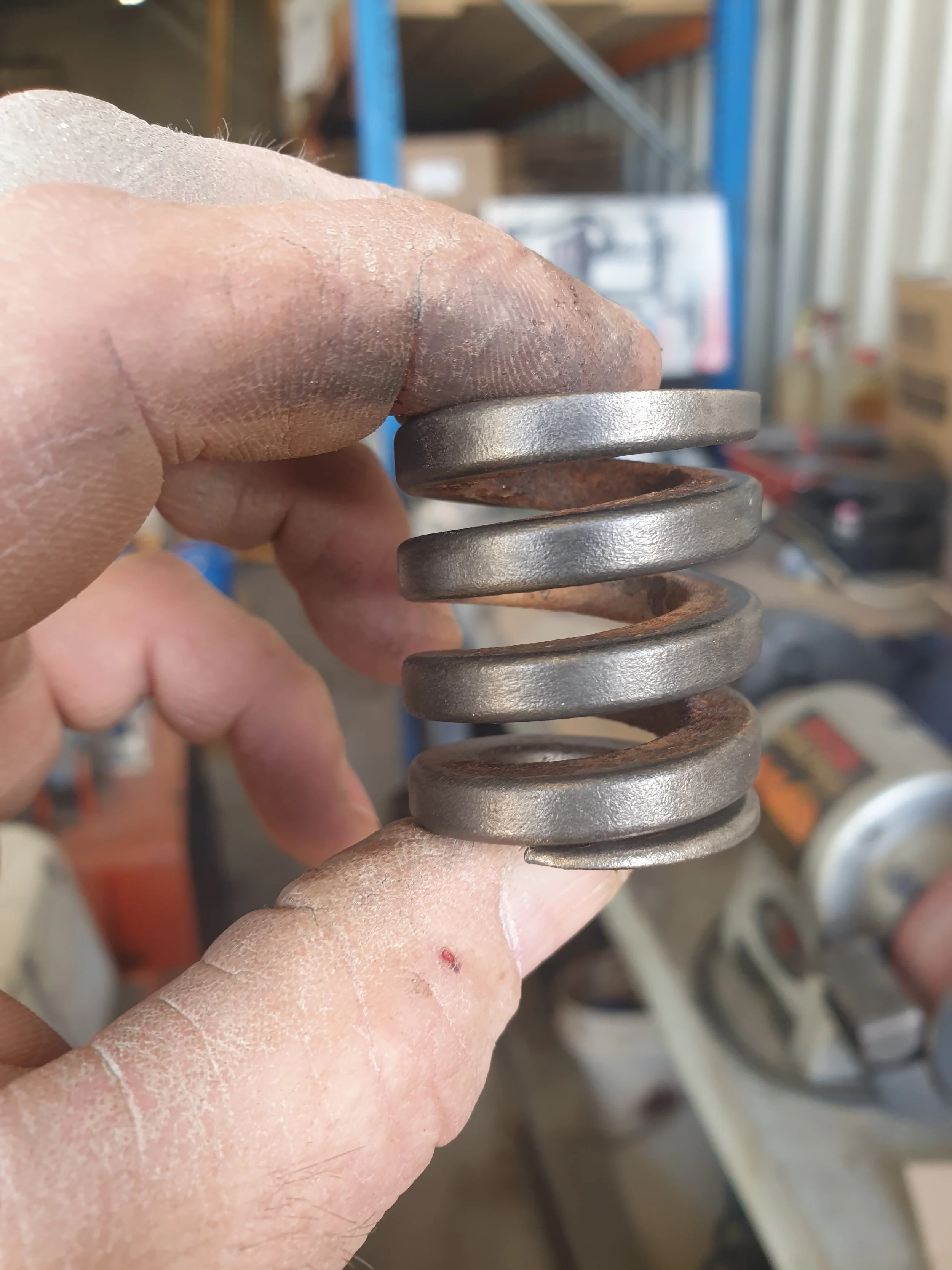 A person's hand holding a rusty metal coil spring with some fresh metal components on top, in a workshop or garage setting.