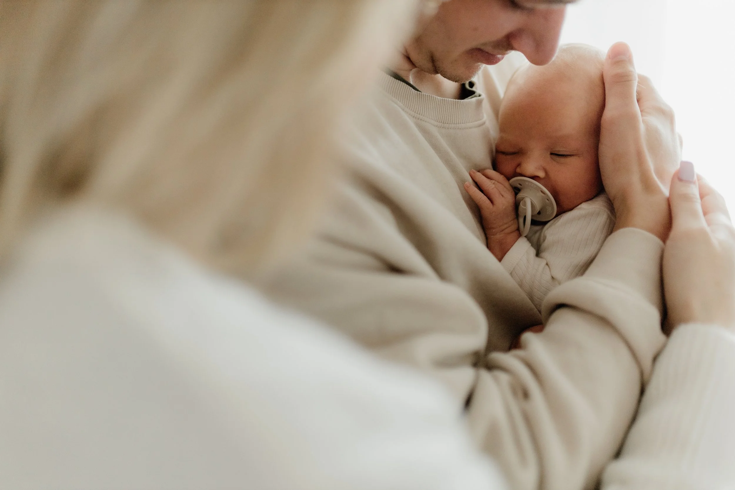 Ein Baby mit Schnuller wird von einem Erwachsenen gehalten und gestreichelt, in einer warmen, hellen Umgebung.