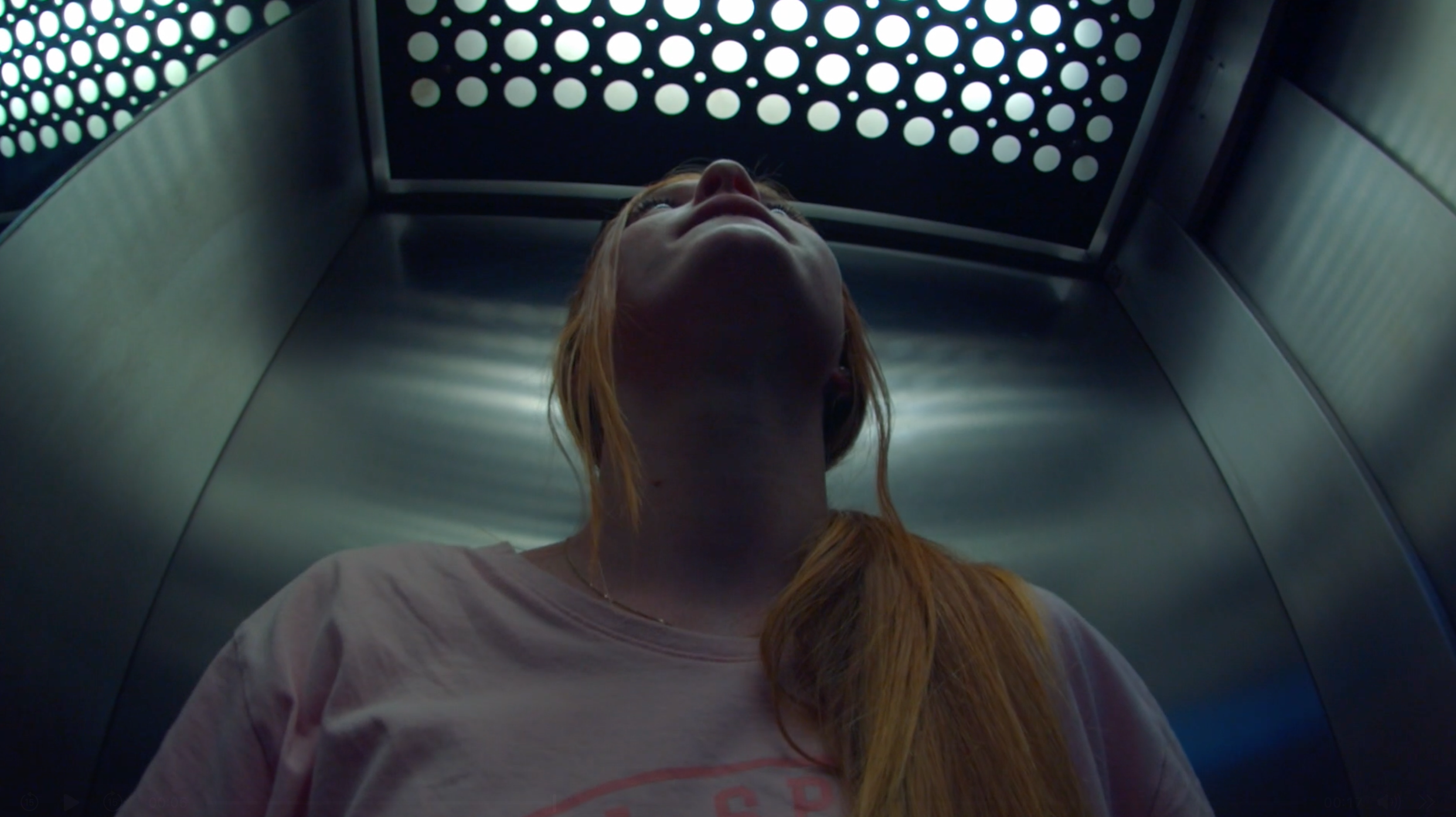 Upward view of a person with long hair in a metallic elevator, looking at the perforated ceiling.