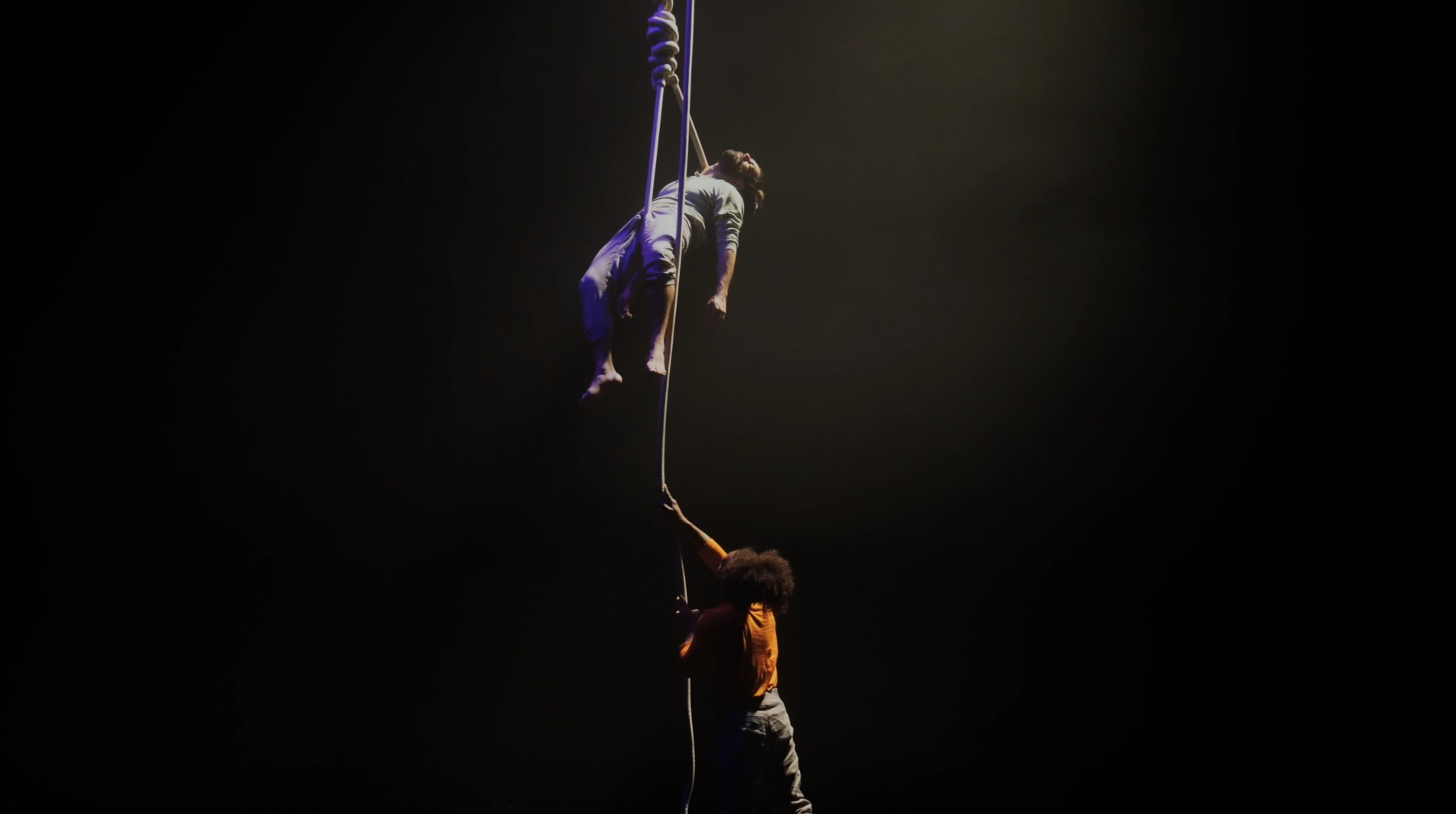 Two performers on a vertically hanging rope in a dimly lit space, with one performer hanging higher and the other reaching up.