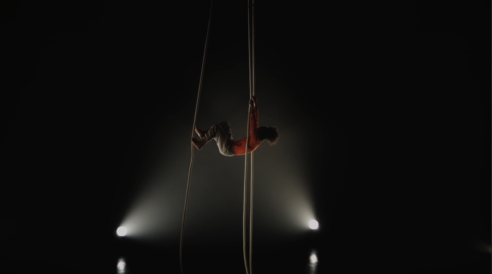 Performer hanging from ropes in spotlight on a dark stage.