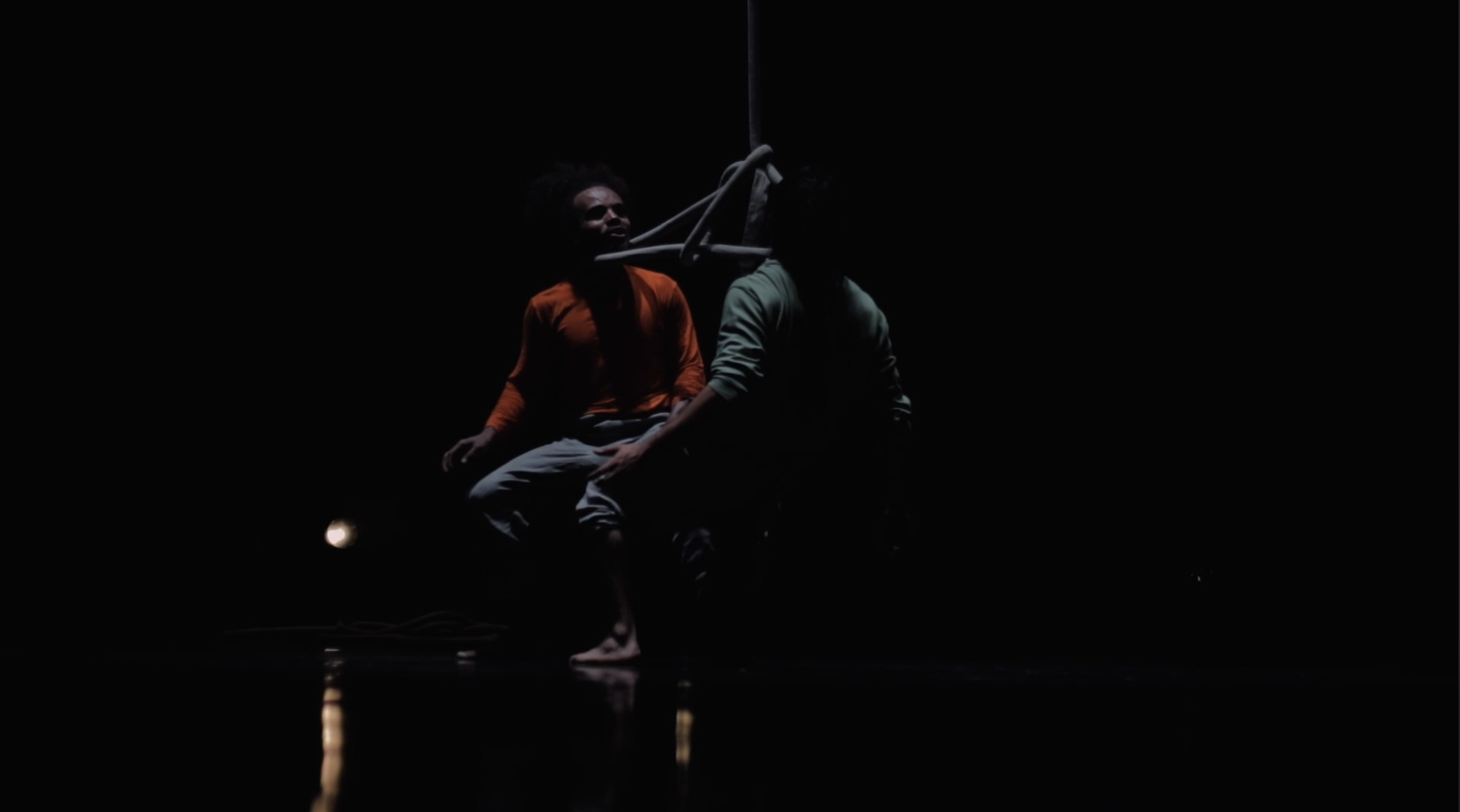A dimly lit scene with two people engaged in aerial acrobatics on a dark stage, one wearing an orange shirt and the other in a light-colored shirt, with ropes or apparatus suspended above.