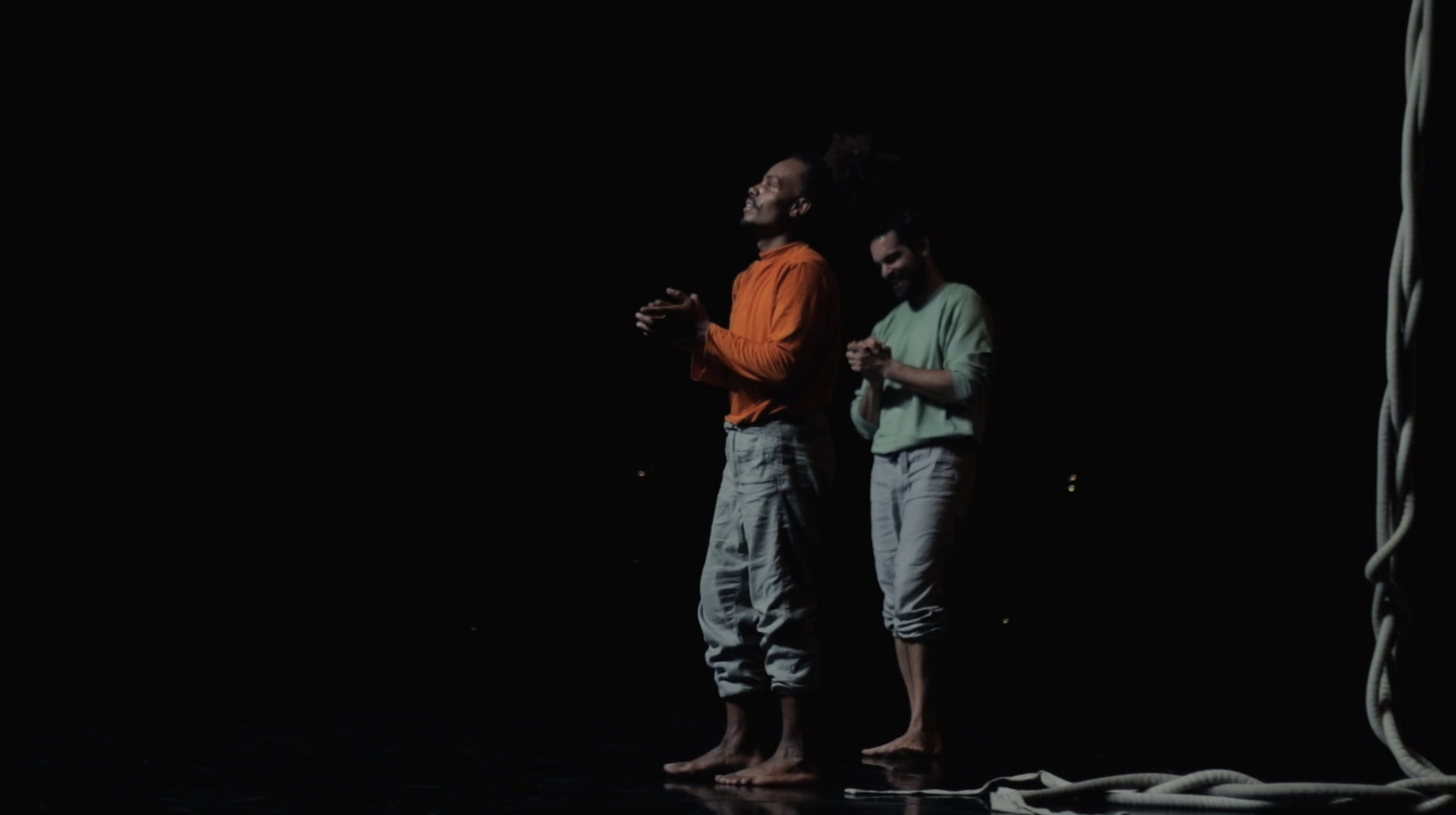Two people standing barefoot on a stage, one wearing an orange shirt and light pants, the other wearing a light green shirt and rolled-up pants. There is a thick rope on the right side of the image.