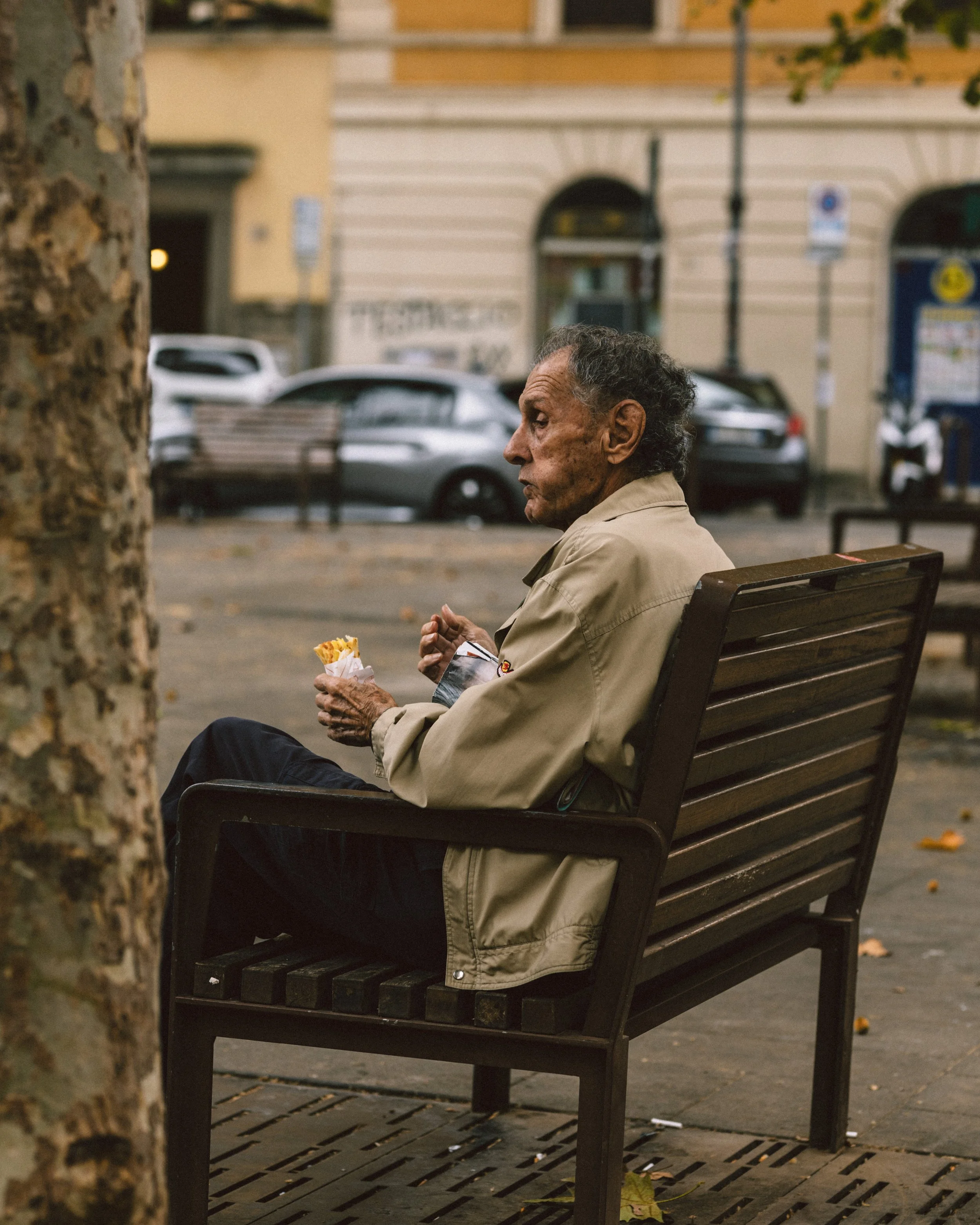 An elderly man sitting on a park bench eating a snack, with cars and buildings in the background.