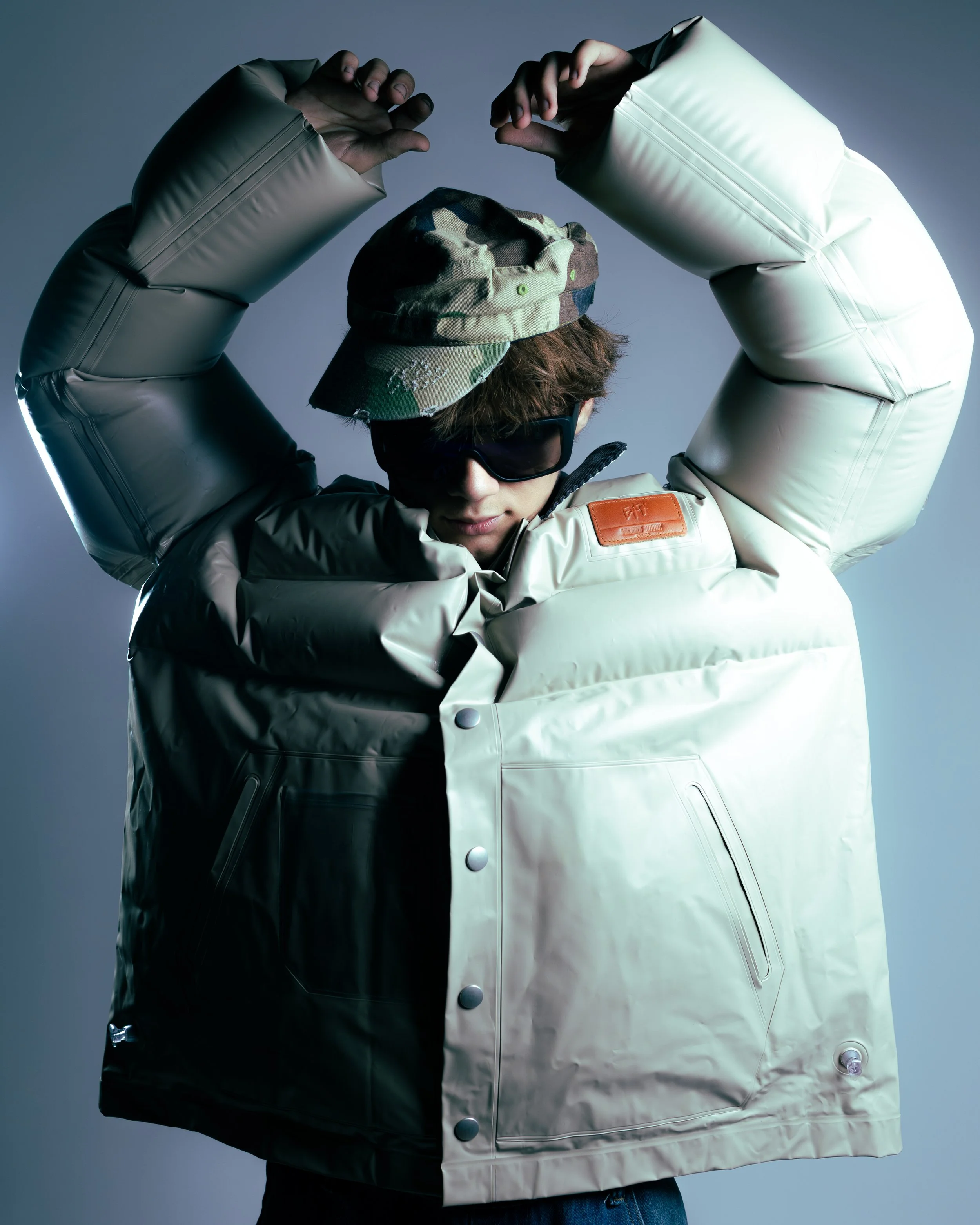 A person wearing a beige puffer jacket, camouflage cap, and sunglasses, posing with arms raised in a flexing position against a neutral background.