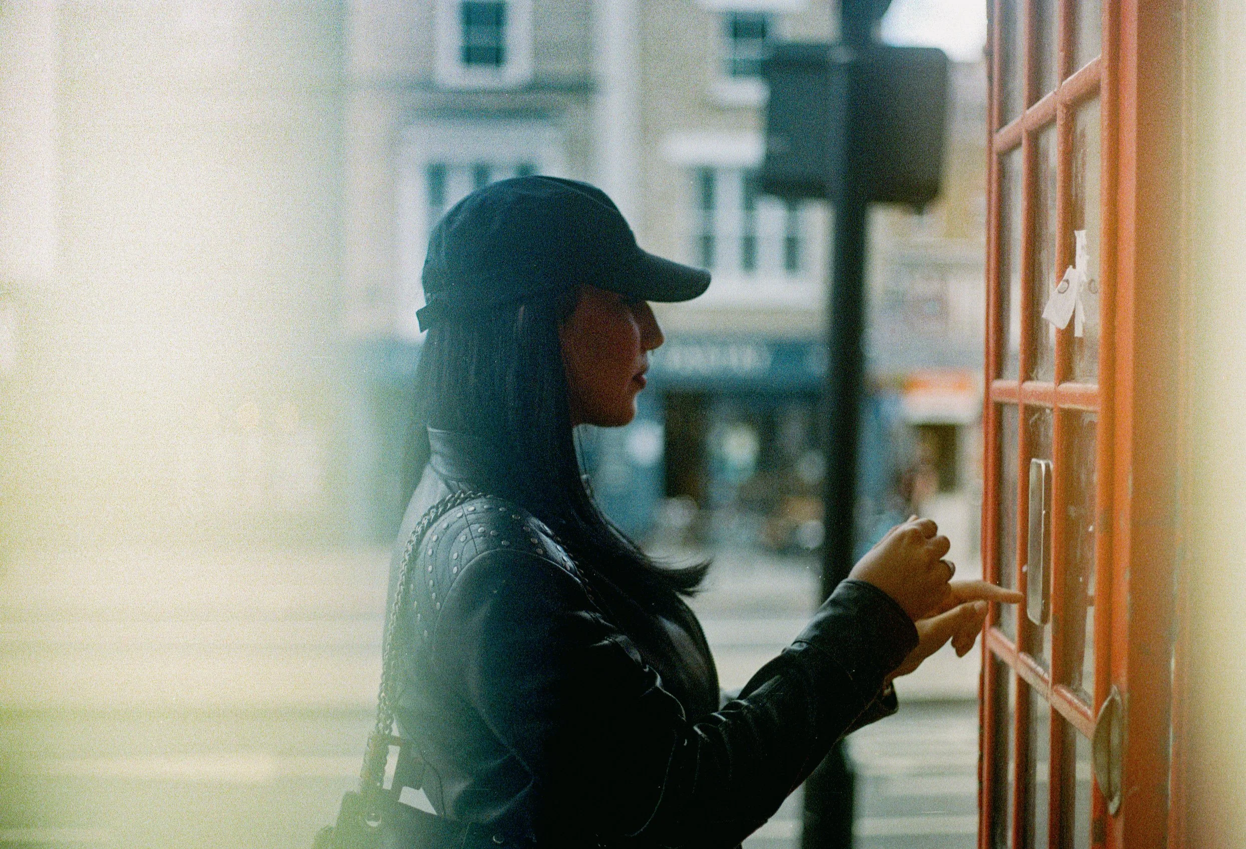A woman wearing a black cap and leather jacket is standing outside, reaching into a red mailbox.