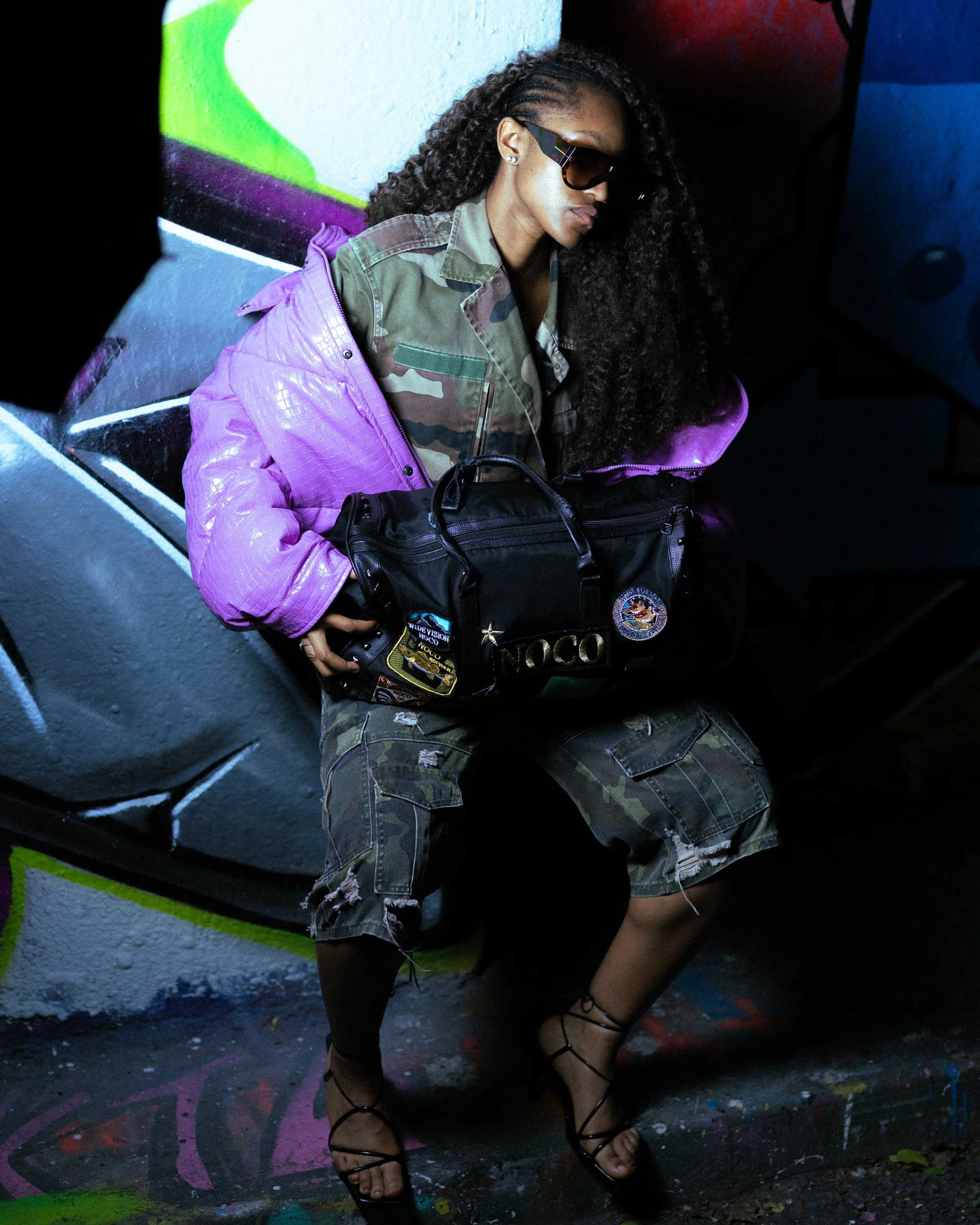A woman with long curly hair wearing sunglasses, a camouflage shirt, a purple jacket, ripped camouflage shorts, and black heels, sitting against a graffiti-covered wall and holding a black bag.