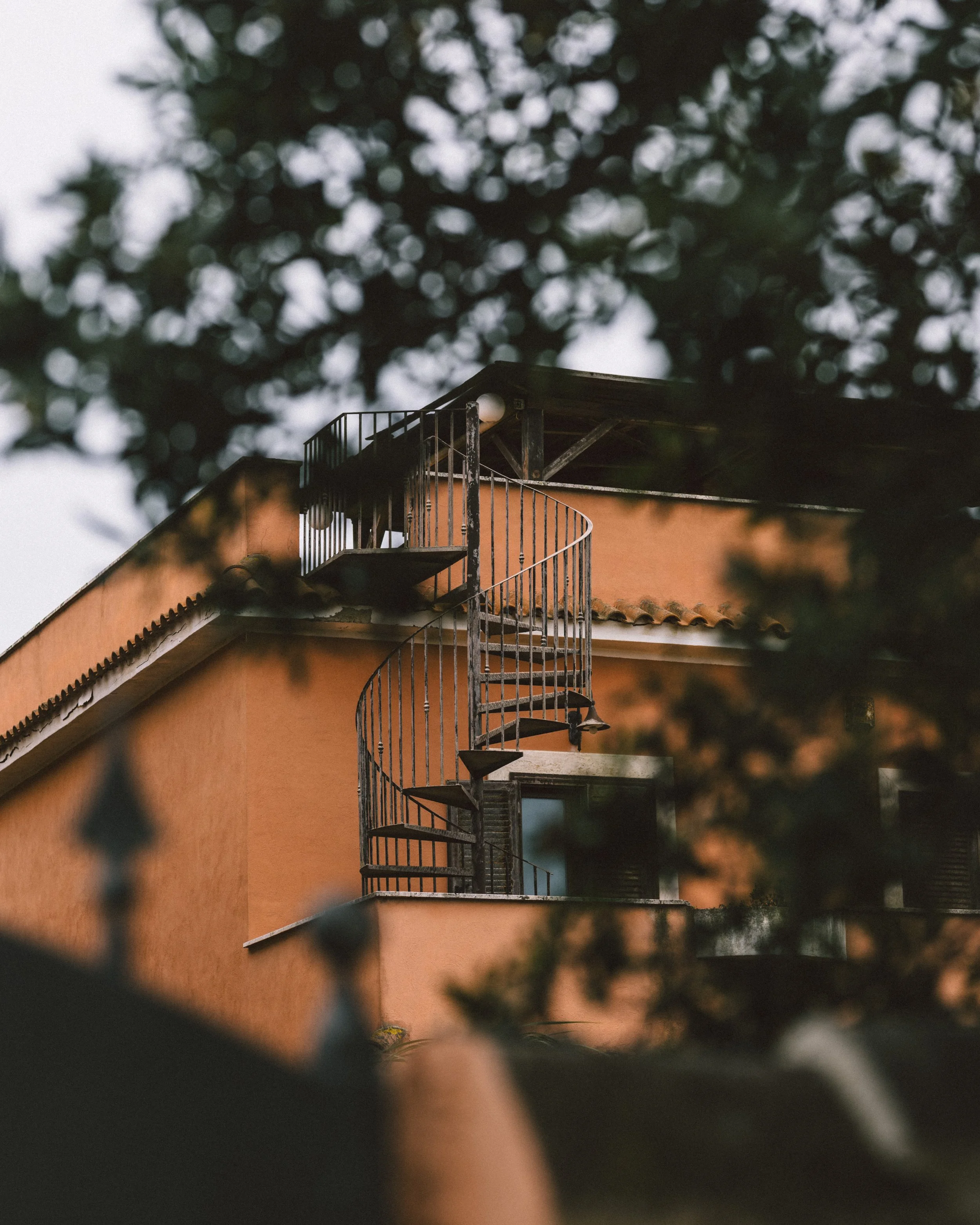 A peach-colored building with an external curly metal staircase, partially obscured by tree branches in the foreground.