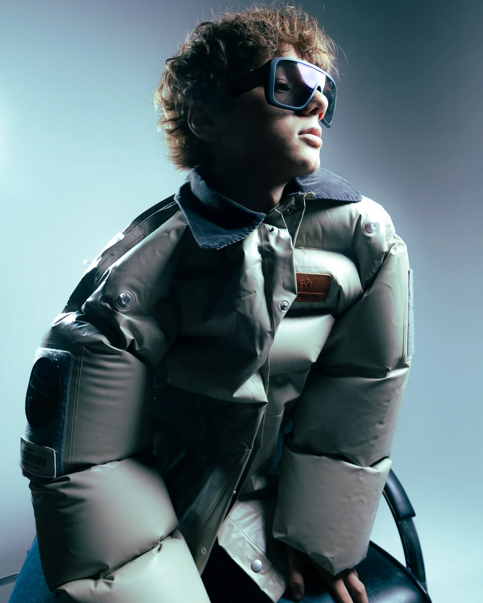 A young man with curly hair wearing a beige puffy jacket and black ski goggles, looking to the side against a plain background.