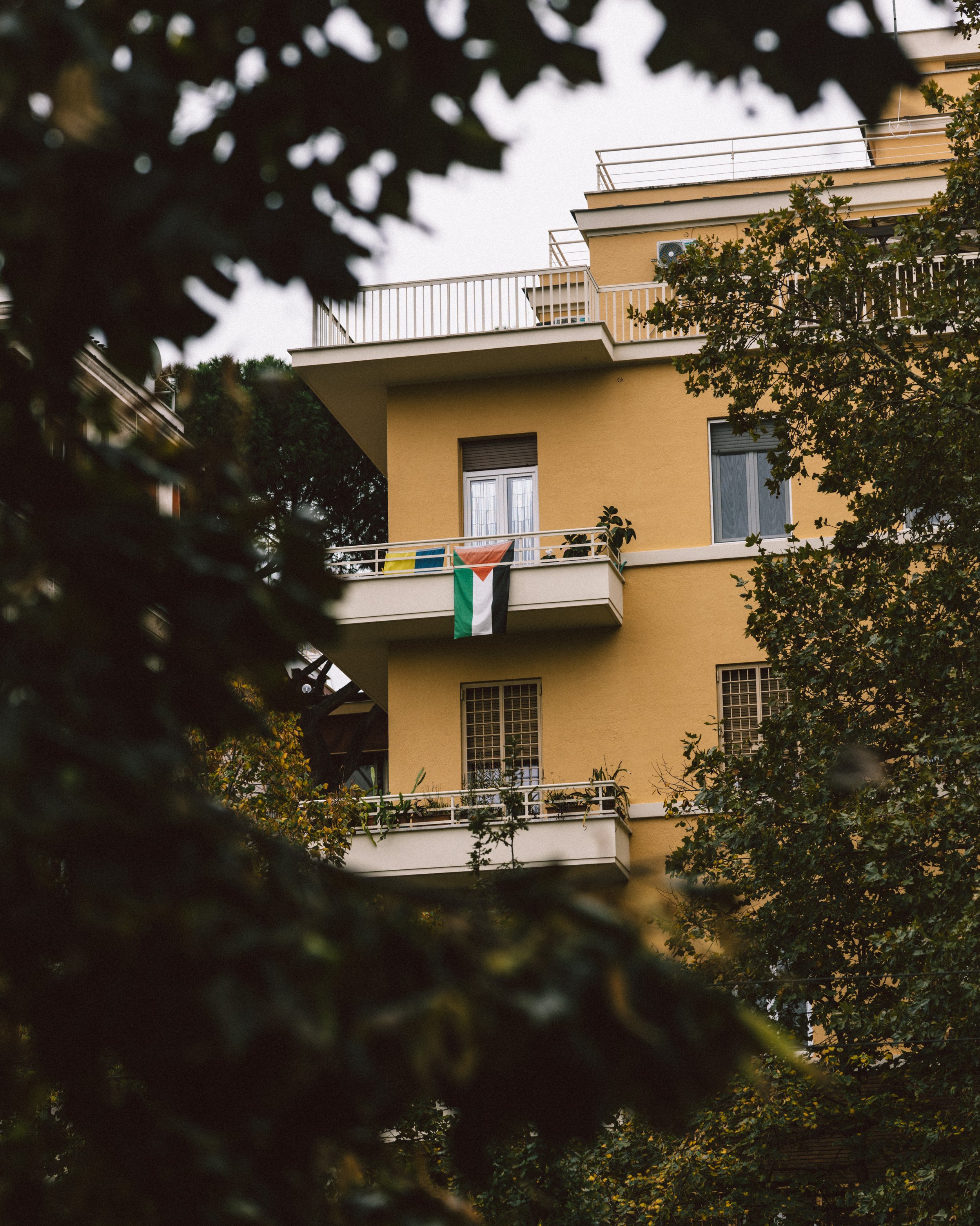 A multi-story yellow apartment building with balconies, framed by tree branches and leaves, with one balcony displaying a flag.