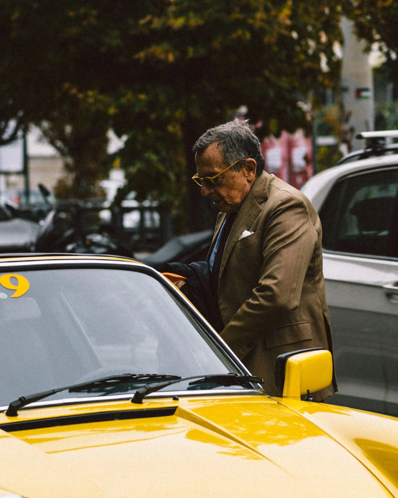 An elderly man in a brown suit and glasses looking into a yellow car in an outdoor parking lot. Trees and parked cars are visible in the background.