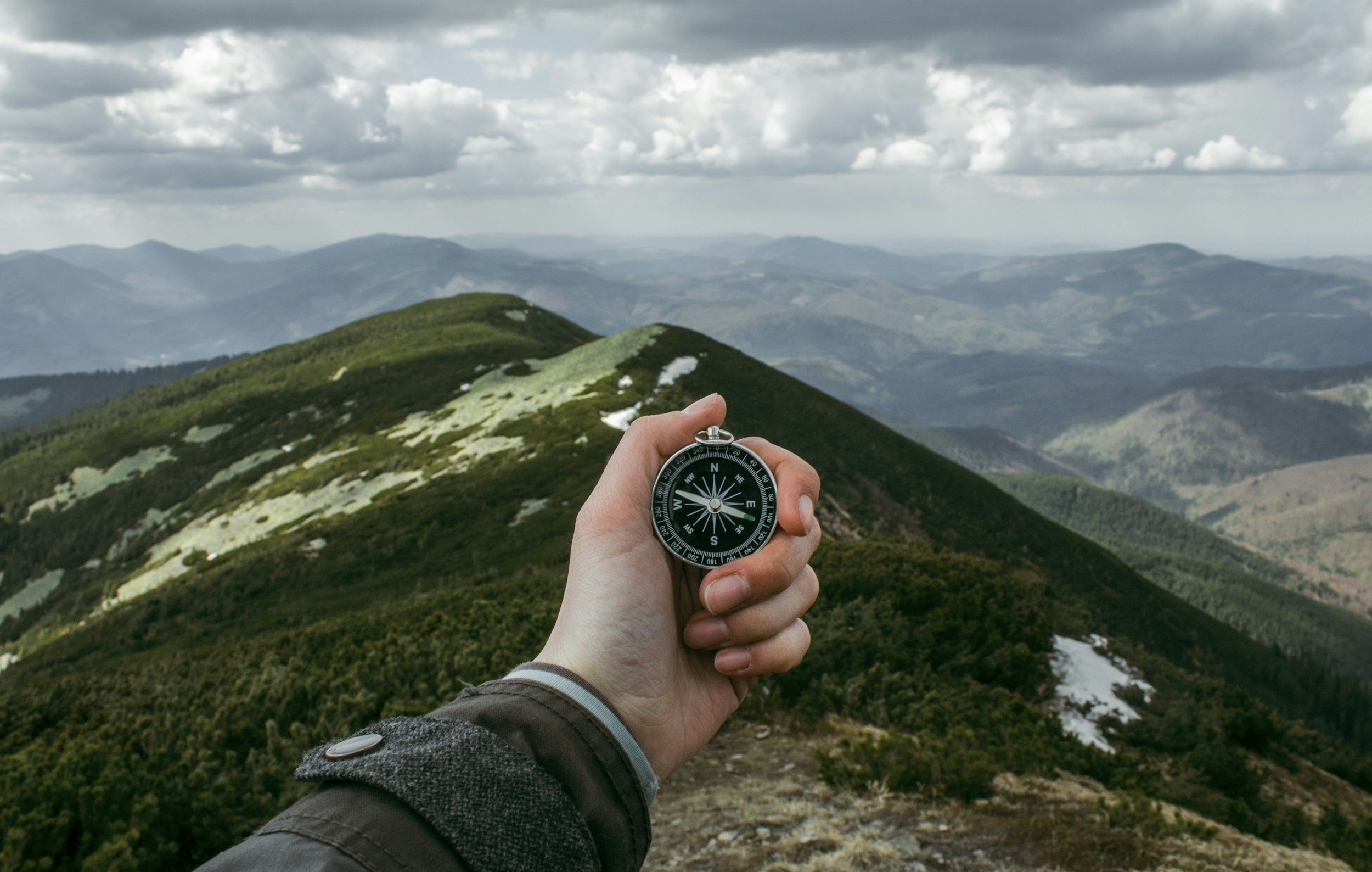 Hand holding a compass with mountainous landscape in the background under cloudy skies.
