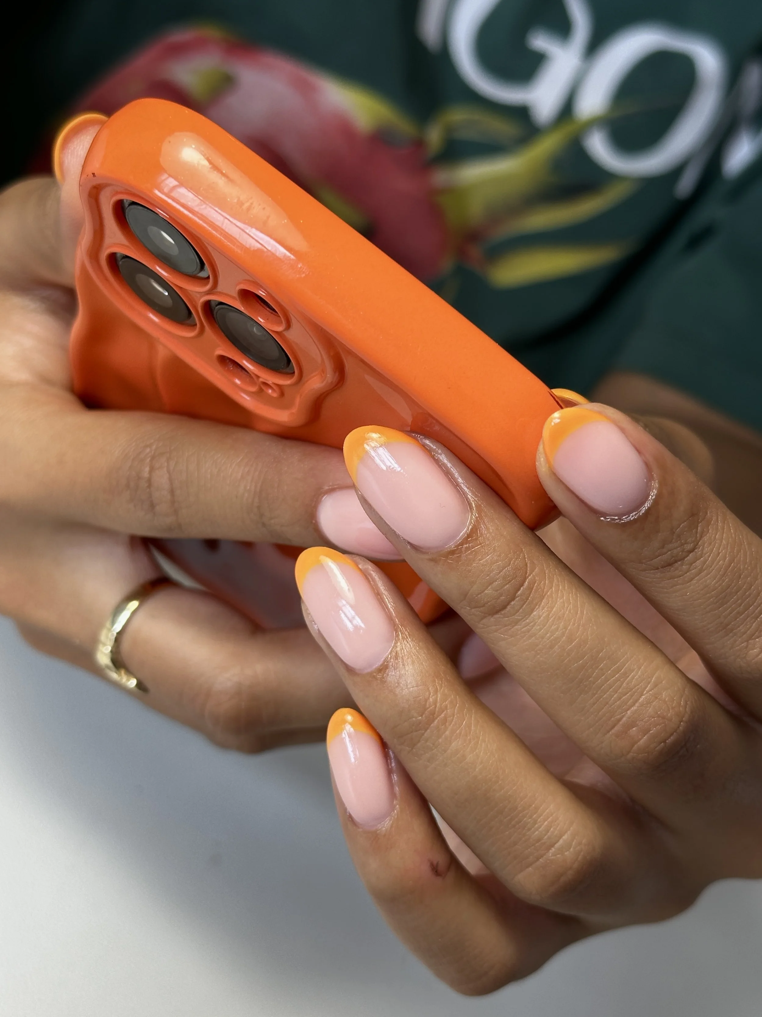 Close-up of a person's hands holding an orange smartphone with a textured back, showing their nails painted with a French manicure style featuring orange tips.