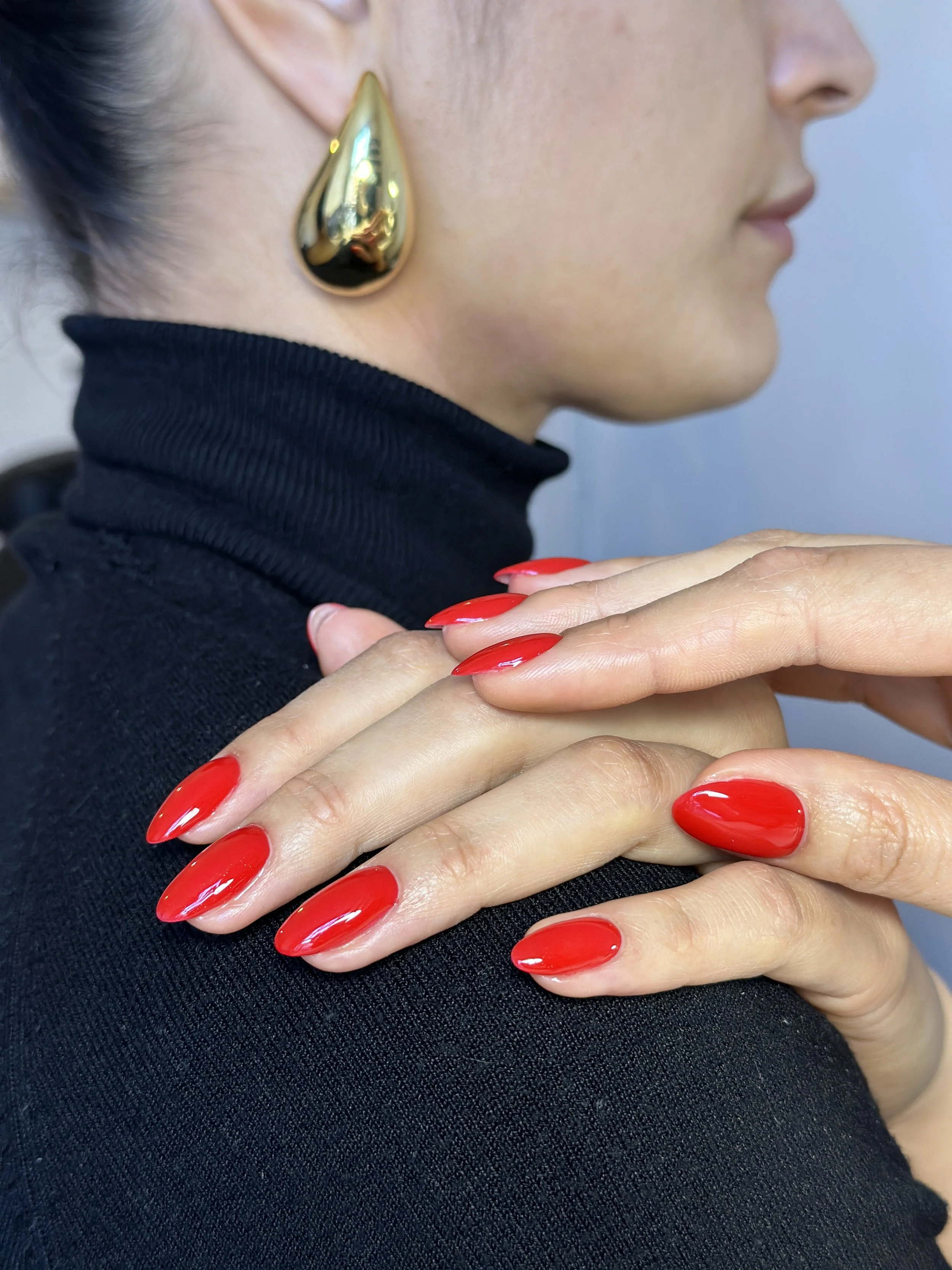 Close-up of a woman wearing a black turtleneck and a large gold teardrop earring, with her hands folded over her shoulder showing bright red nail polish.