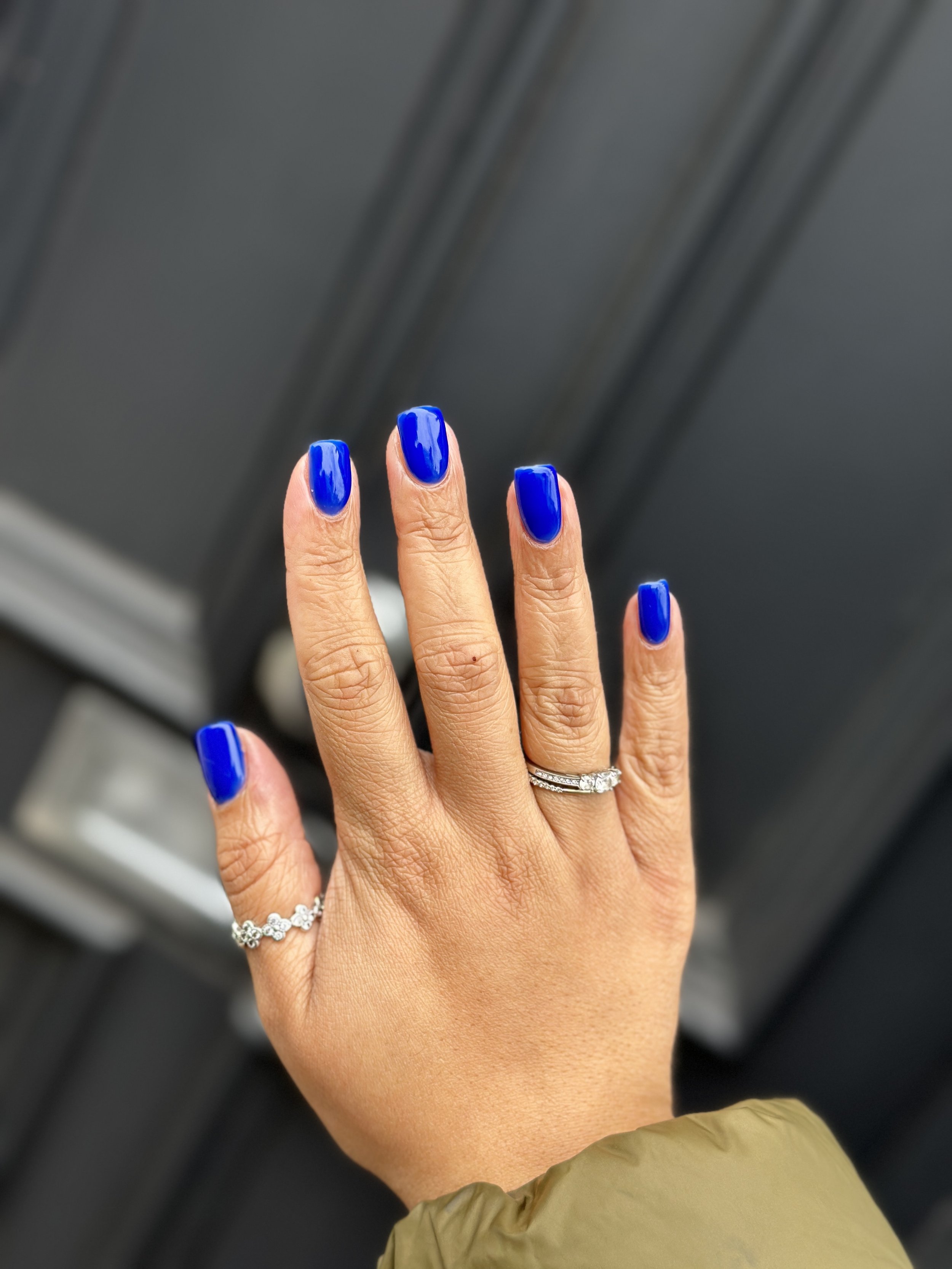 Close-up of a hand with a blue manicure, wearing two rings, held against a dark background.