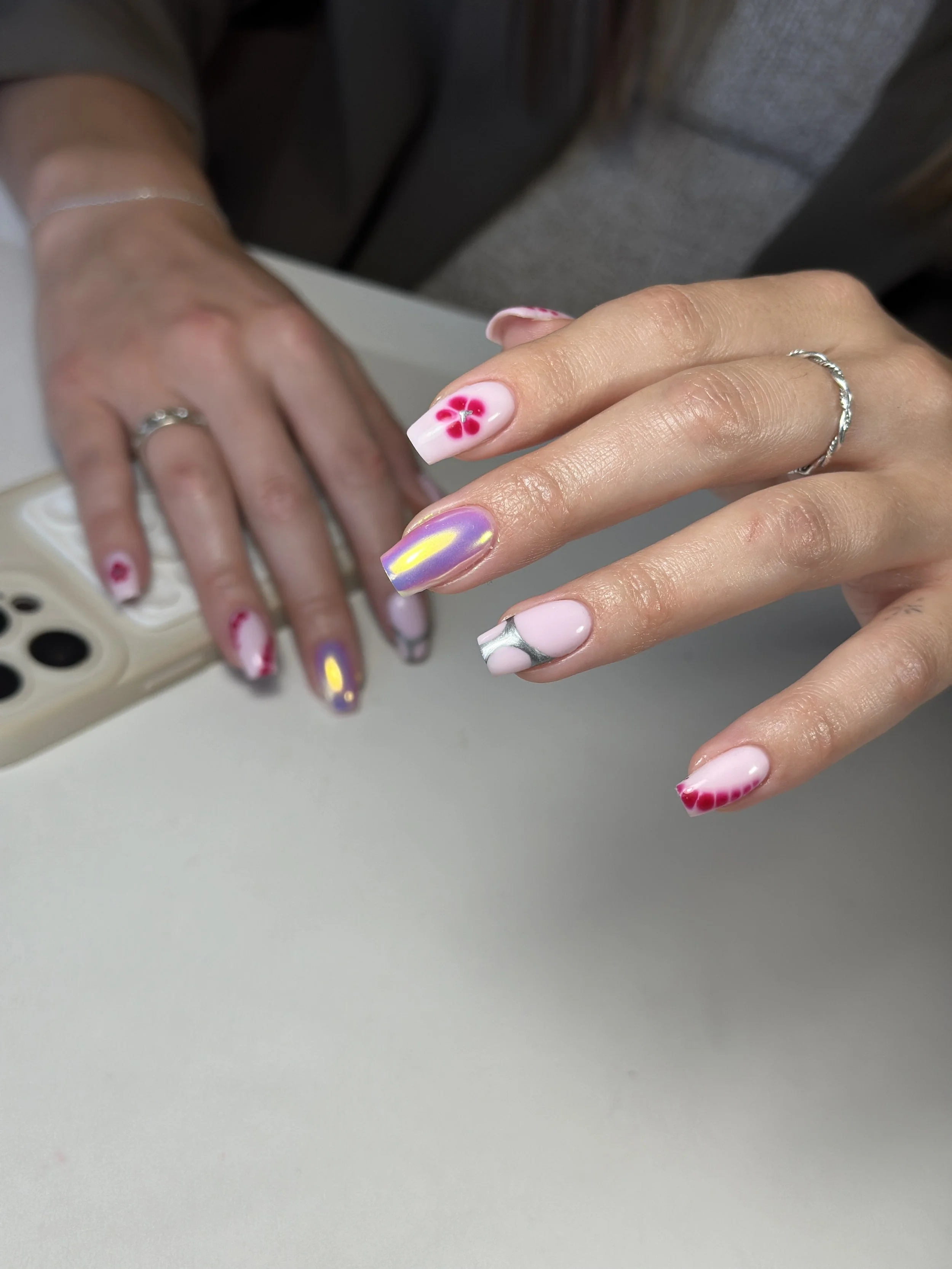 Close-up of hands with colorful floral and iridescent nail art, one hand showing rings, holding a phone with a beige case on a gray surface.