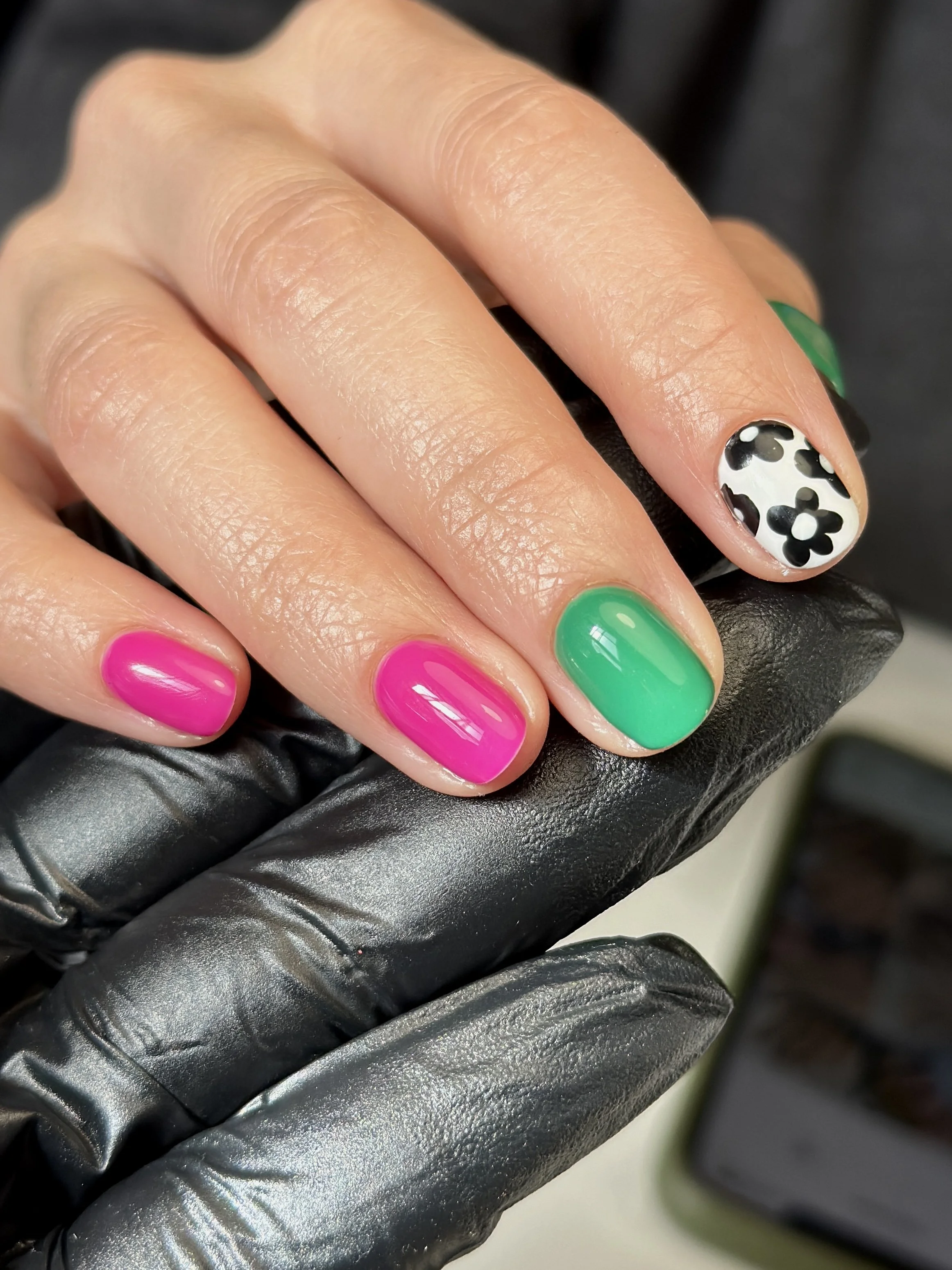 Close-up of a woman's hand with colorful manicured nails, including pink, green, and black-and-white floral designs, resting on a black glove with a blurred background.