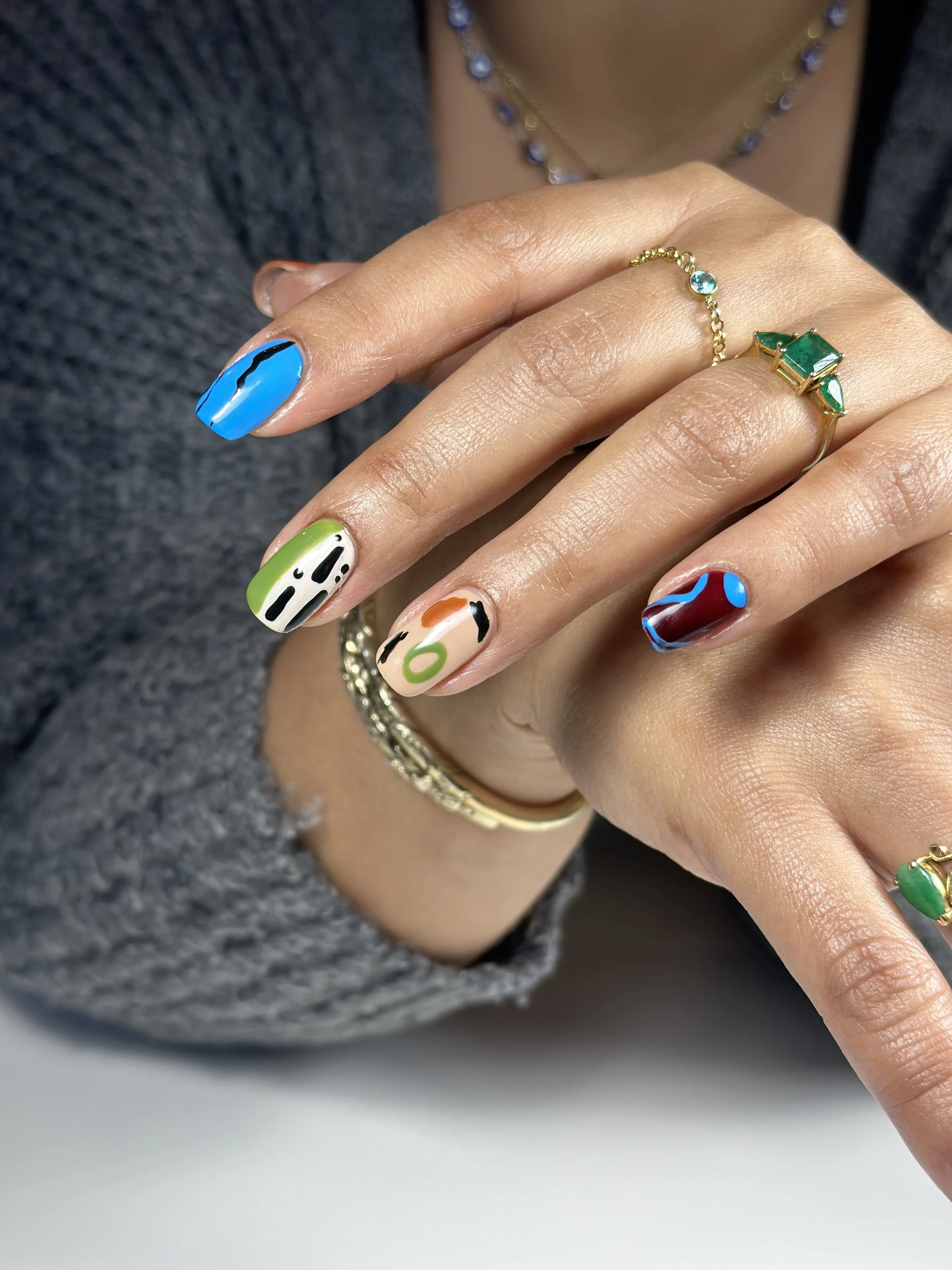 Close-up of a woman's hand with colorful abstract nail art, multiple gold rings, and a delicate gold and blue beaded necklace.