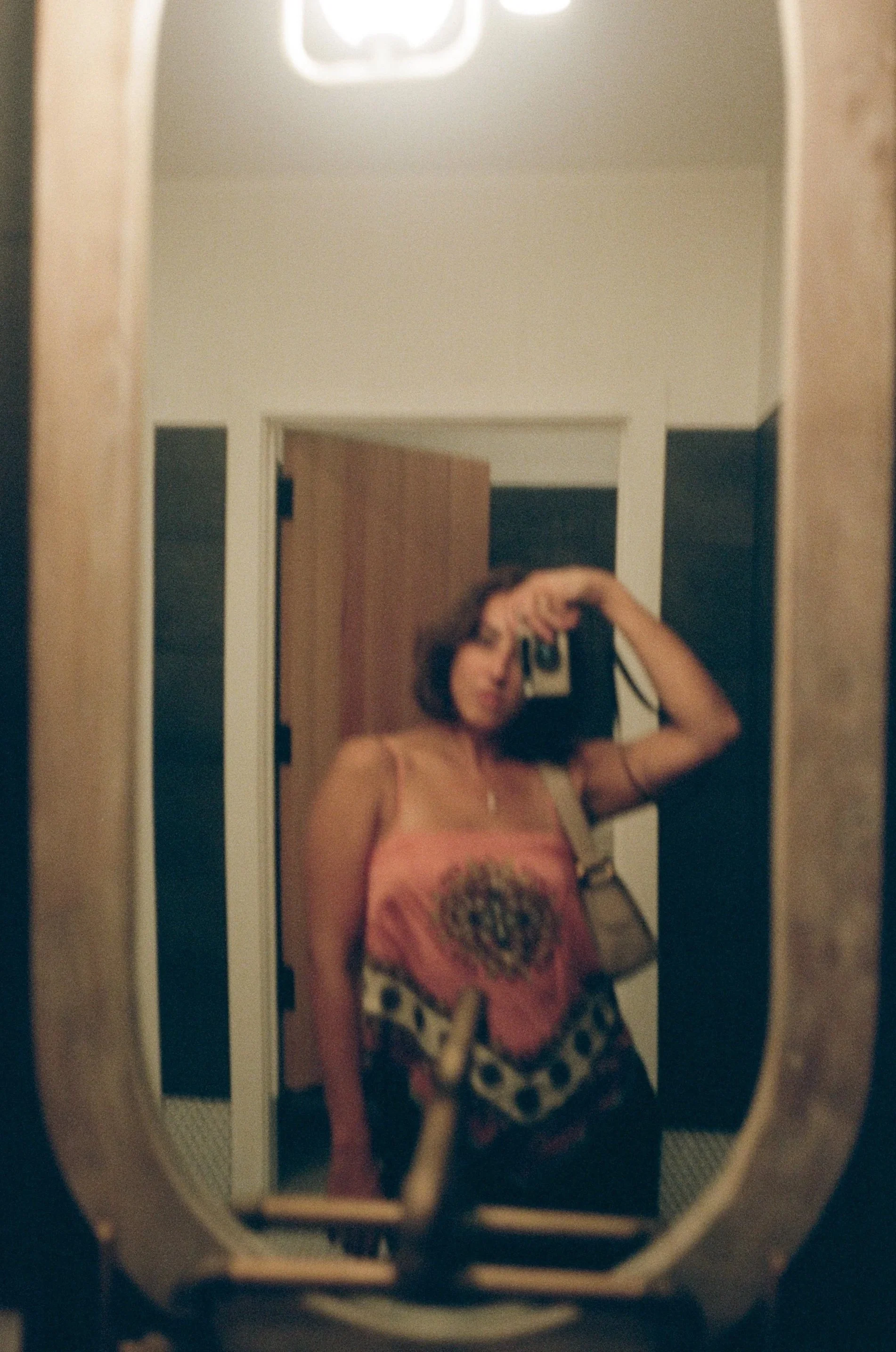 Wider mirror self-portrait of Emily Zoe standing with a camera raised, framed by a curved mirror and softly lit interior.