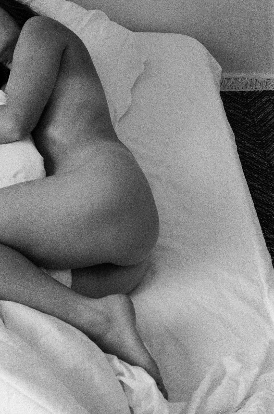 Black-and-white analog photograph of a nude body curled on white bedding, focused on form, gesture, and light.