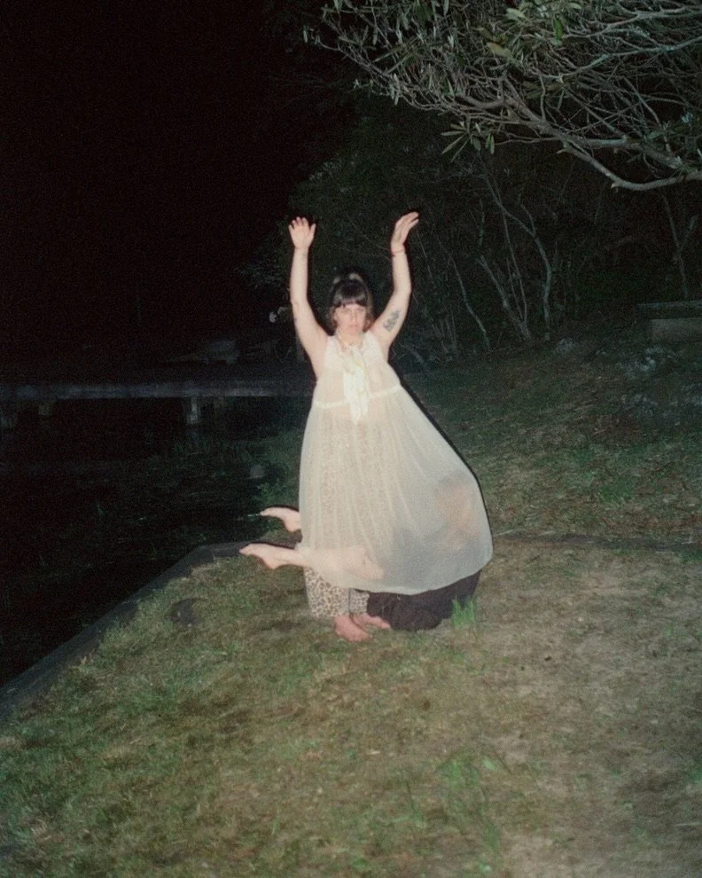 Flash photograph of a woman in a pale dress raising her arms outdoors at night.