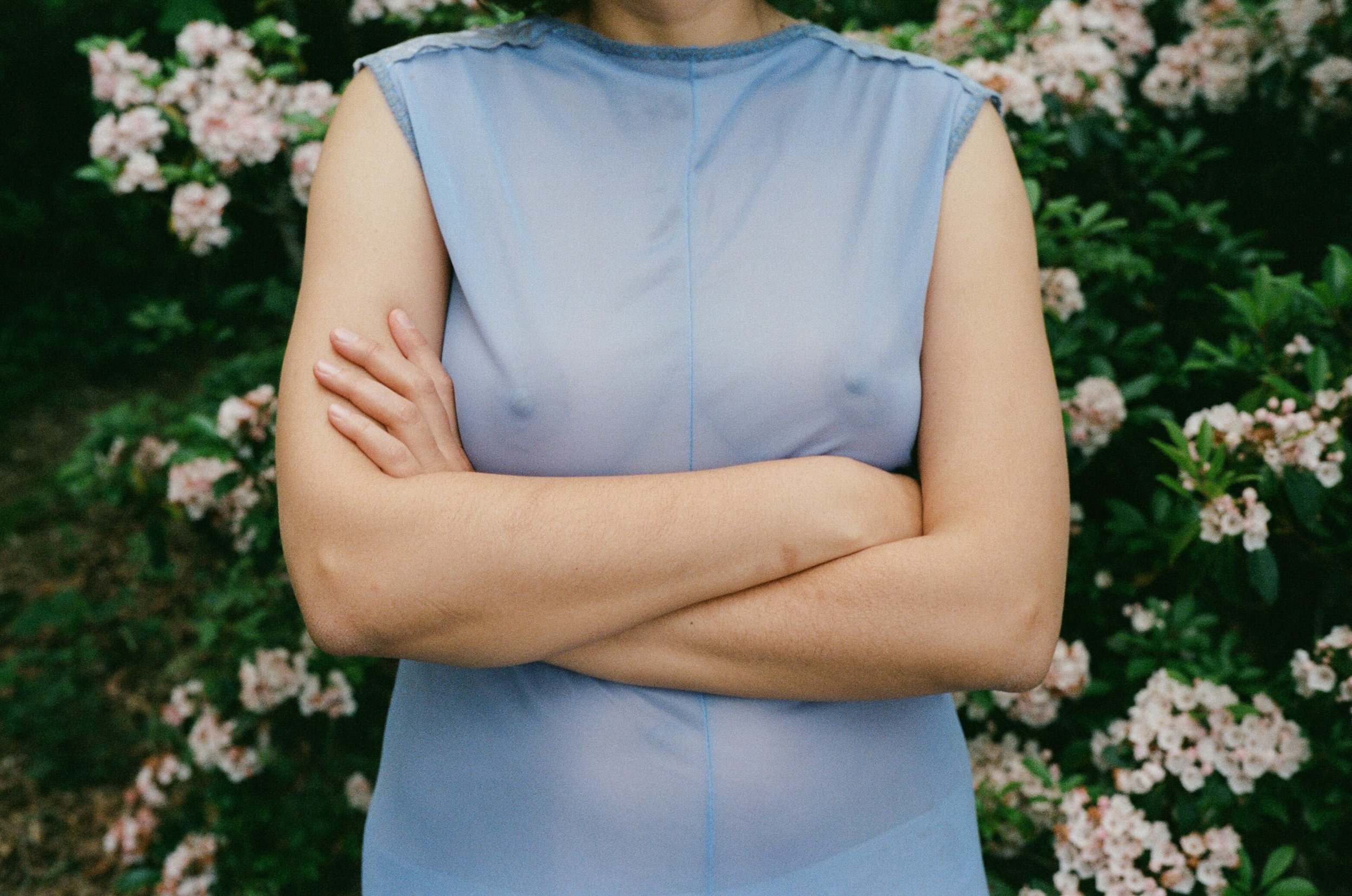 Film portrait of a woman in a pale blue dress standing in front of flowering bushes.