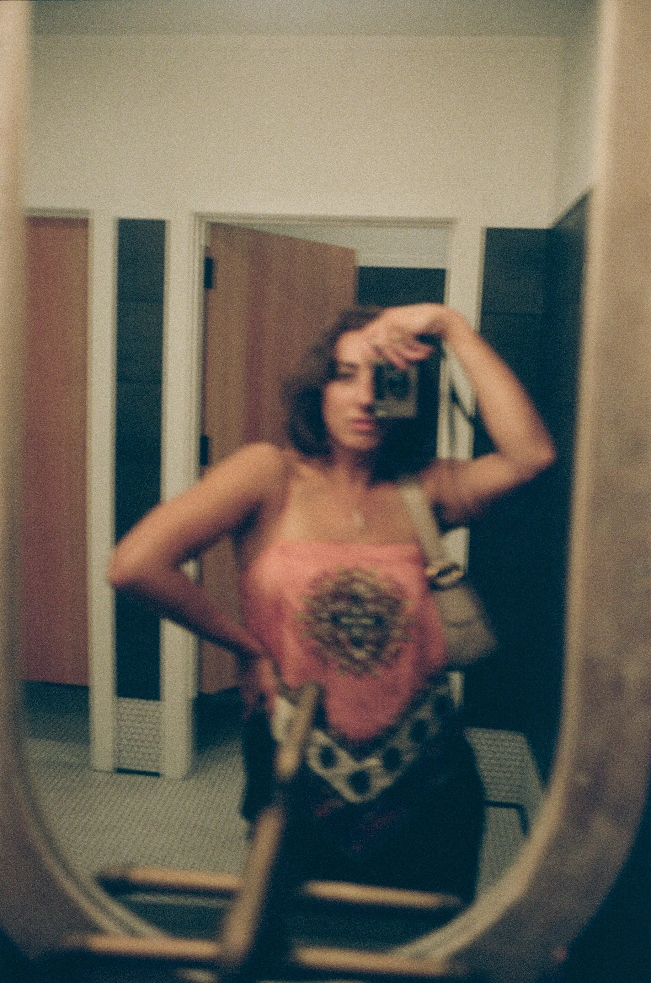 Soft-focus mirror self-portrait of artist Emily Zōe holding a camera above her face, wearing a pink top in a warmly lit interior.