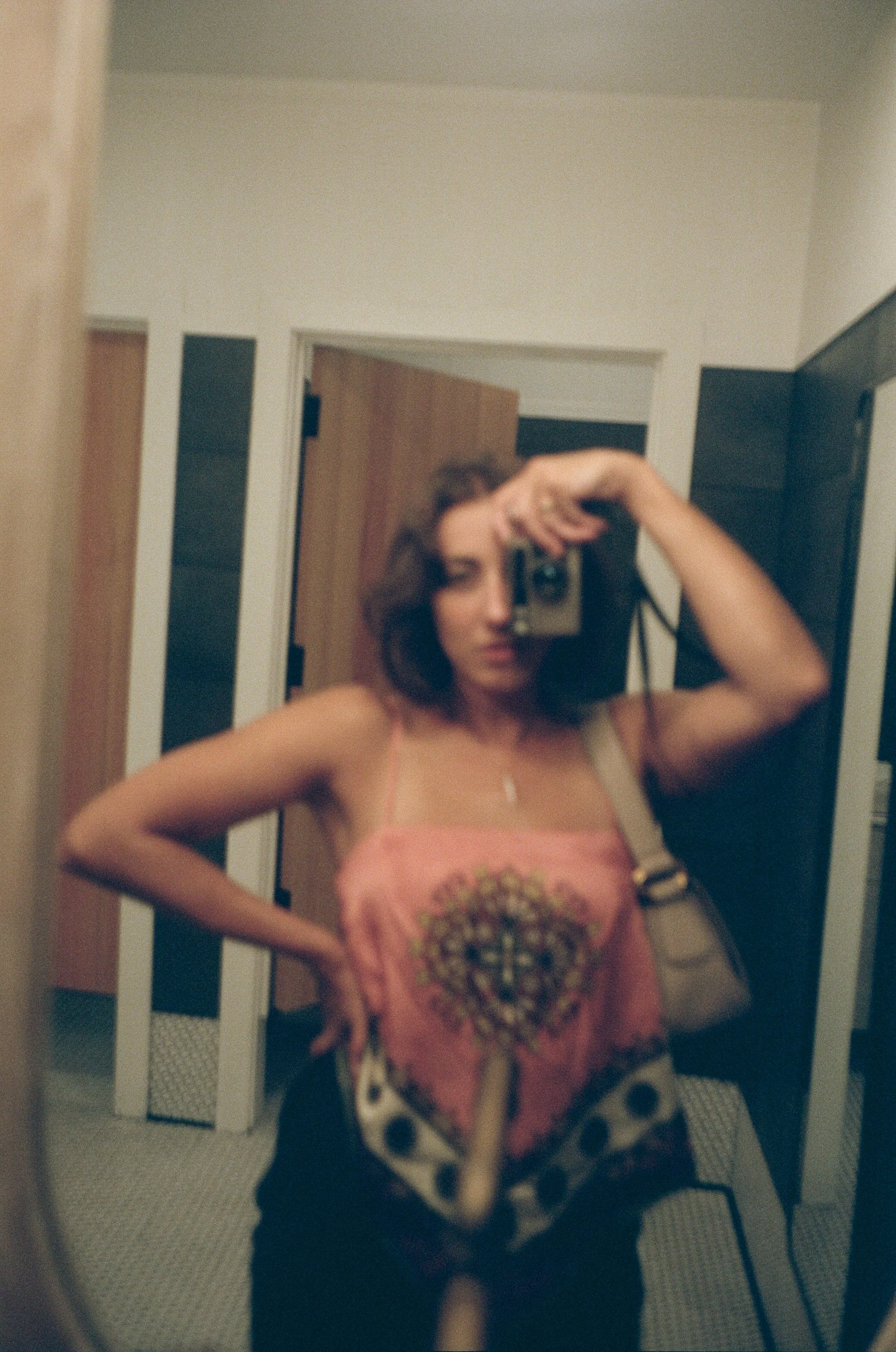Soft, grainy self-portrait of Emily Zoe with one hand resting on her hip and a camera raised to her face, reflected in a mirror.