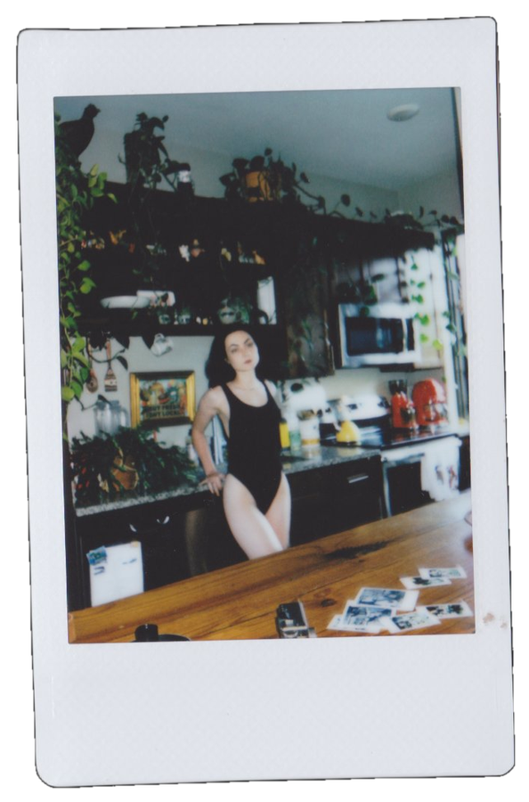 A woman wearing a black swimsuit standing in a kitchen next to a wooden table with scattered instant photos, kitchen appliances, and plants.