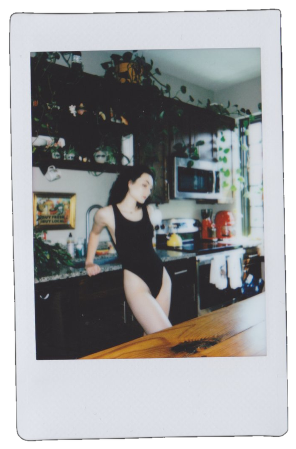 A woman in a black swimsuit standing in a kitchen with a countertop, microwave, and various kitchen items, with greenery and patterned curtains in the background.