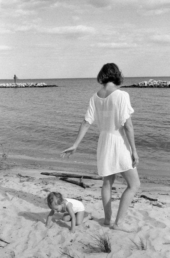 Black-and-white film photograph of a woman in a white dress standing with a young child on a beach.