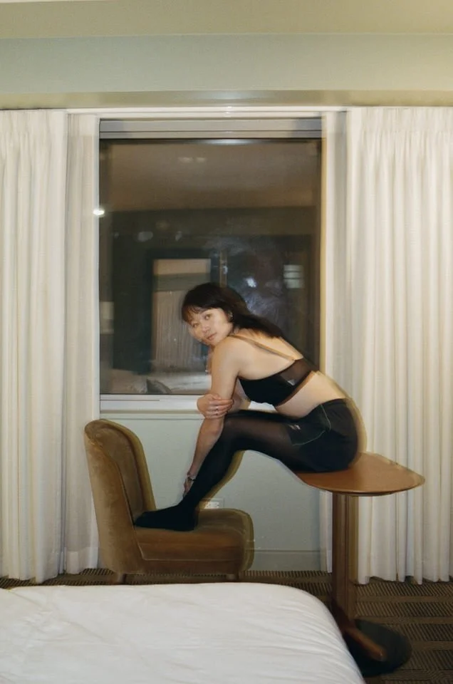 Analog film portrait of a woman in black clothing perched between a chair and table in a softly lit hotel room.
