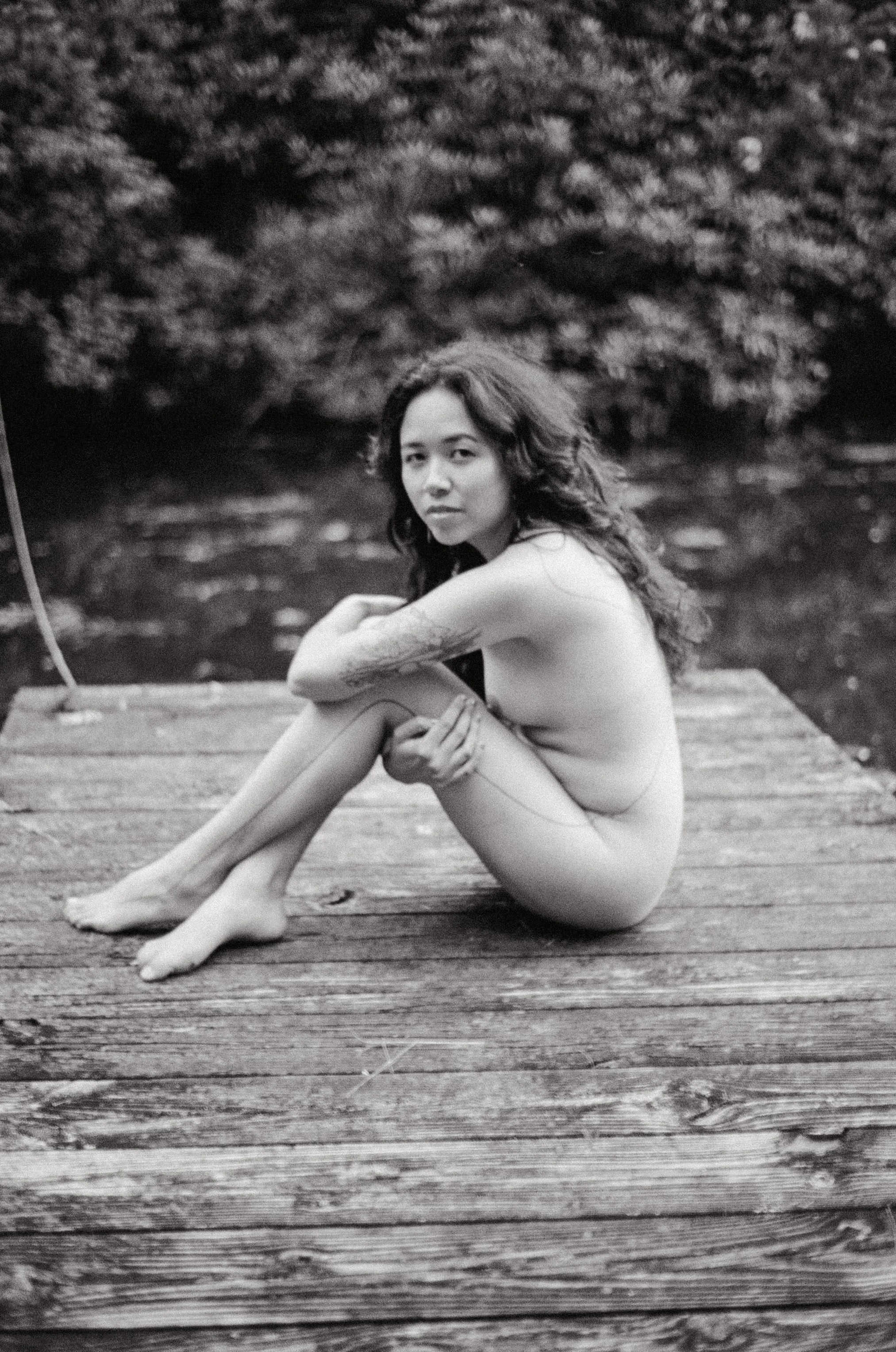 Black-and-white film portrait of a nude woman seated on a wooden dock beside the water.