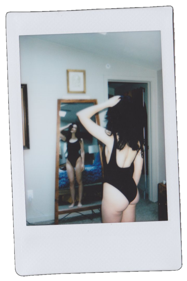 A woman in a black swimsuit standing in front of a mirror, taking a photo of her reflection, in a bedroom with framed artwork on the wall.