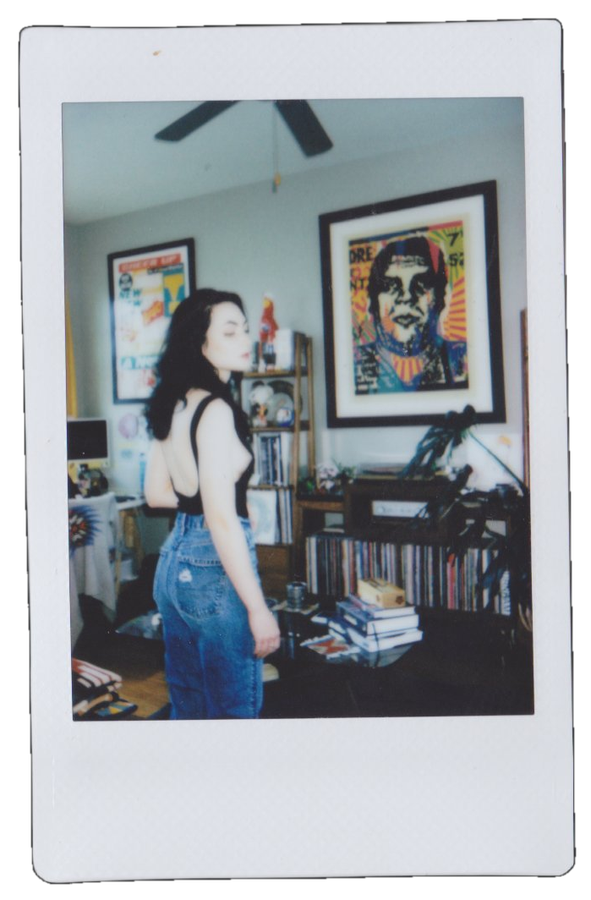 A woman with black hair wearing a black top and high-waisted jeans, standing indoors in a room with framed artwork on the wall, books, and electronic equipment on shelves and tables.