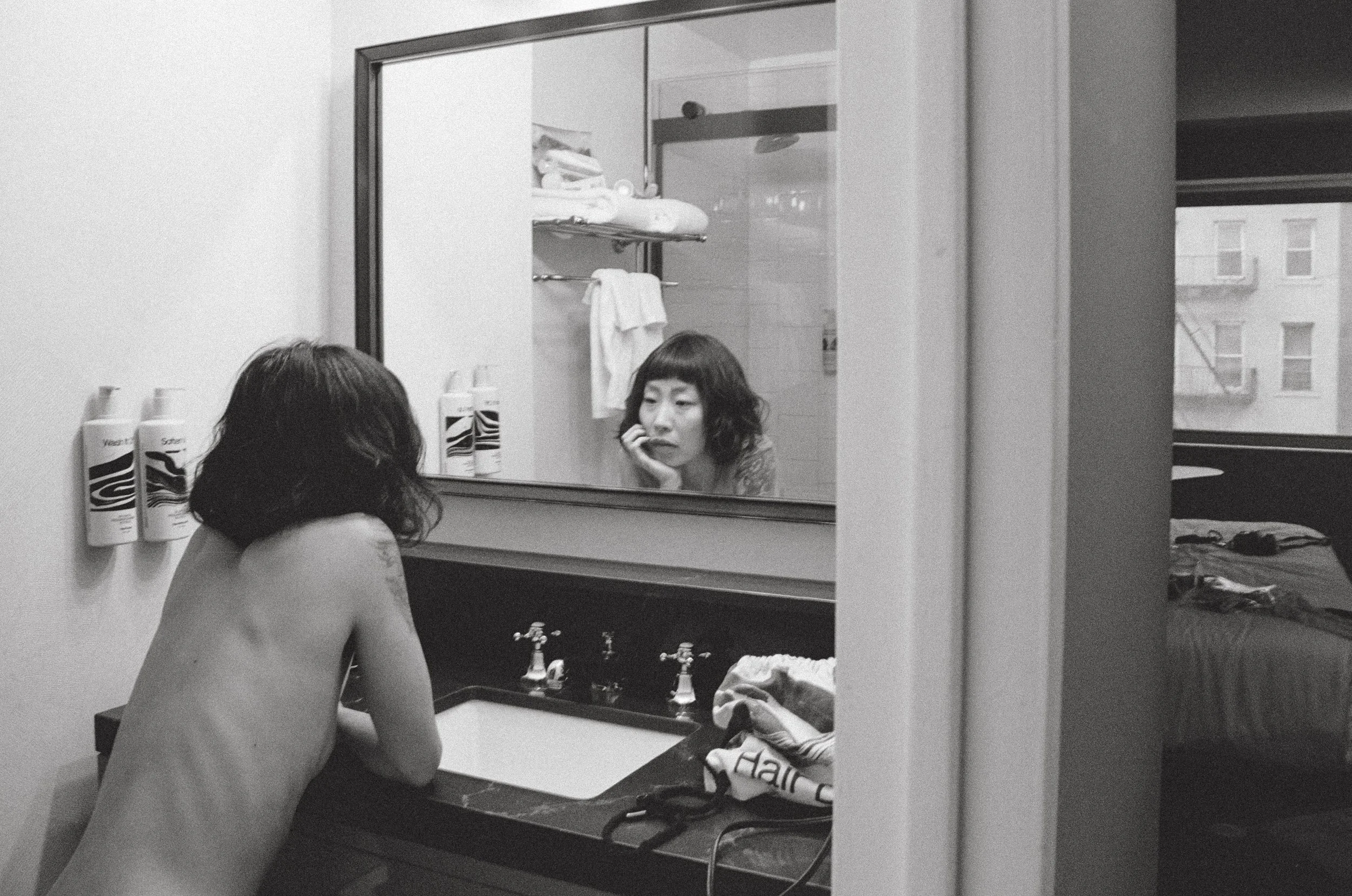 Black-and-white analog portrait of a woman seen from behind and reflected in a bathroom mirror.
