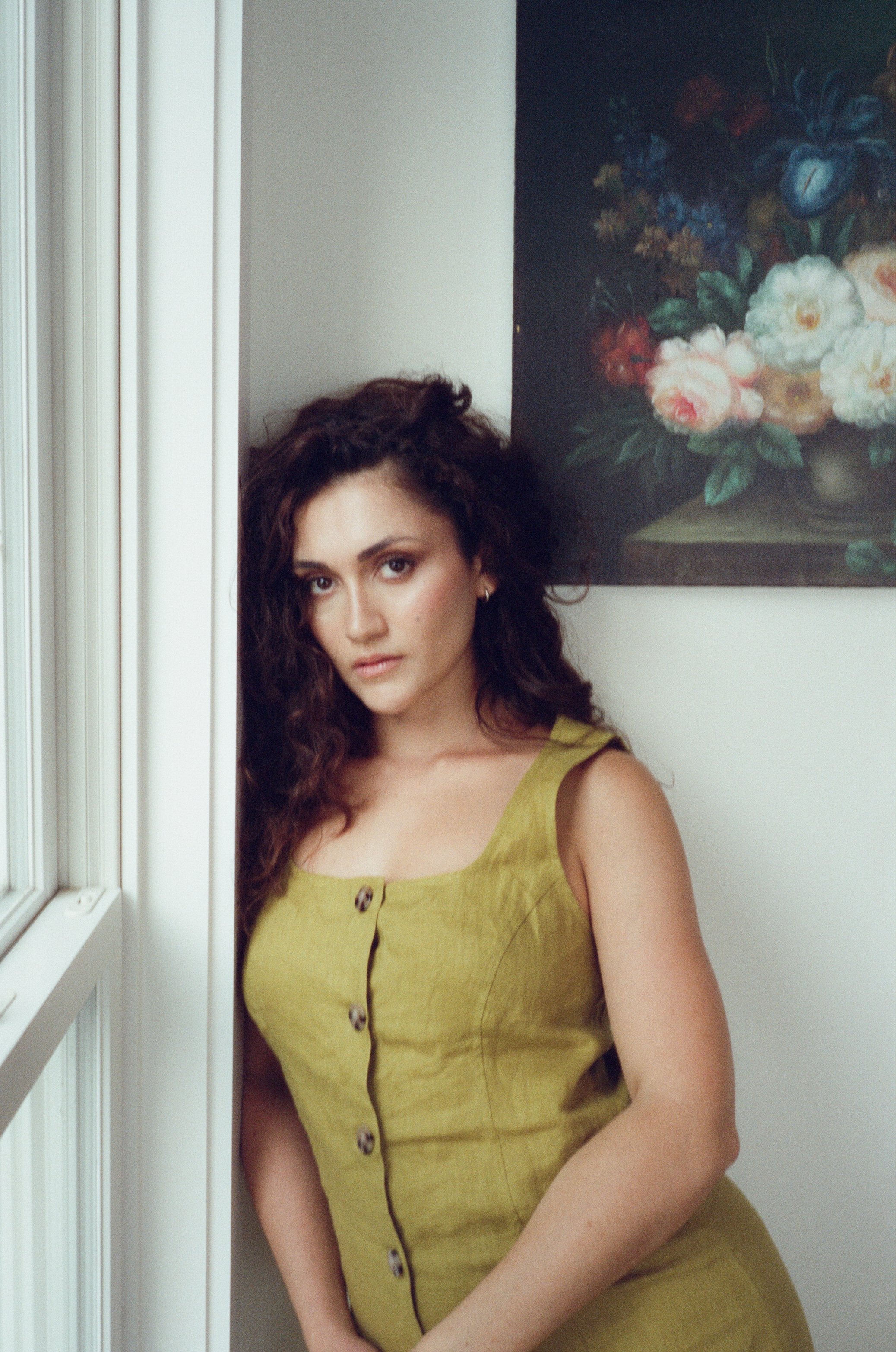 Analog film portrait of a woman with dark curly hair wearing olive green beside a window, photographed by Emily Zoe.