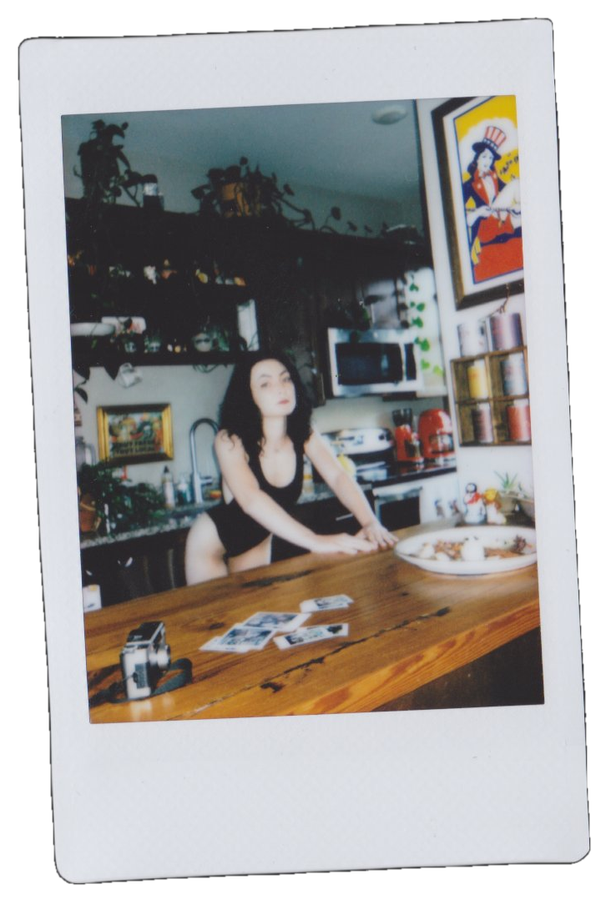 A woman with dark hair in a black outfit leaning on a kitchen counter in a colorful kitchen with artwork and kitchen appliances, with a camera and photographs on the counter in front of her.