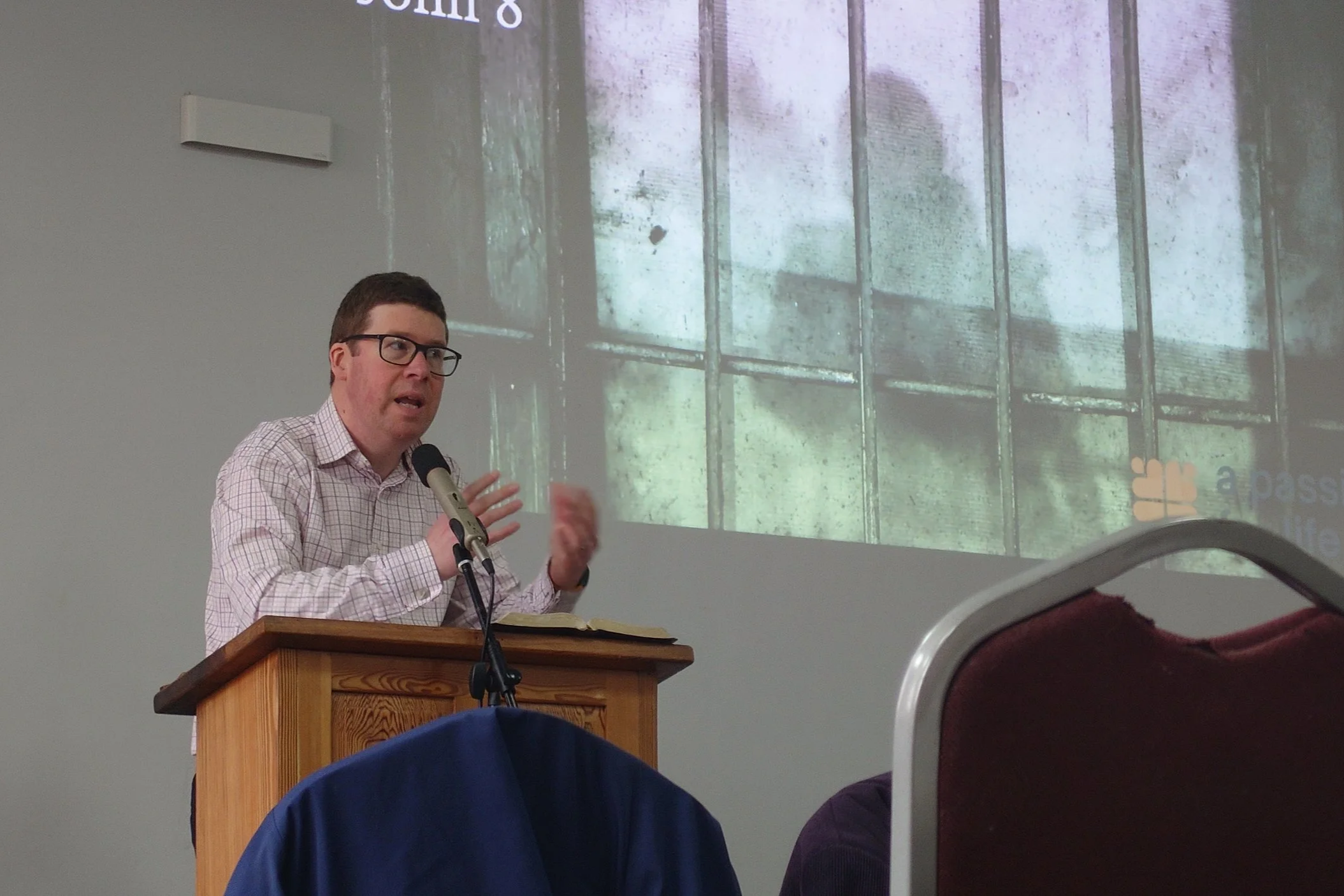 A man with glasses, wearing a checked shirt, is speaking into a microphone behind a wooden podium at a presentation or lecture. A large projected image of a window with bars and grime is visible on the wall behind him.