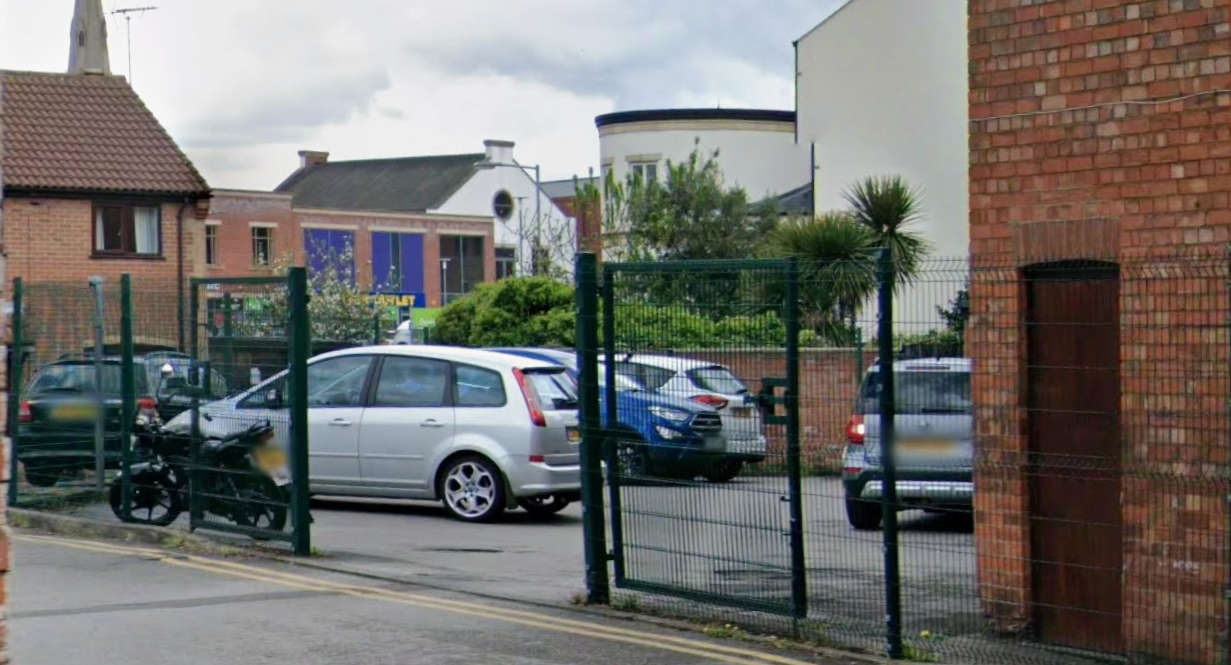 A parking lot with several cars, a motorcycle, and a gate surrounding it, with buildings and greenery in the background.