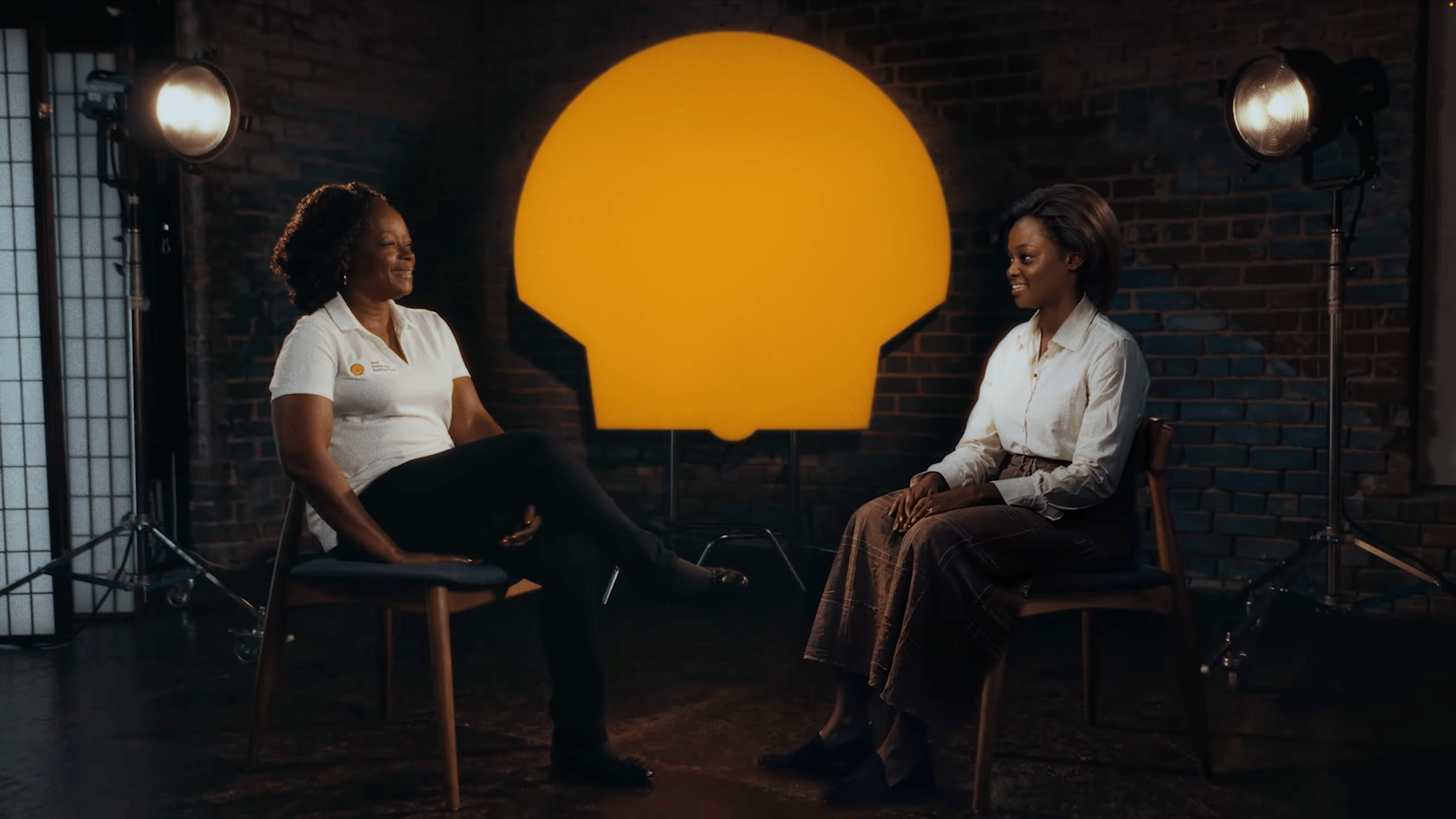 Two women sitting and talking in a studio with a large illuminated yellow lightbulb shape on the wall behind them, brick wall background, studio lighting.